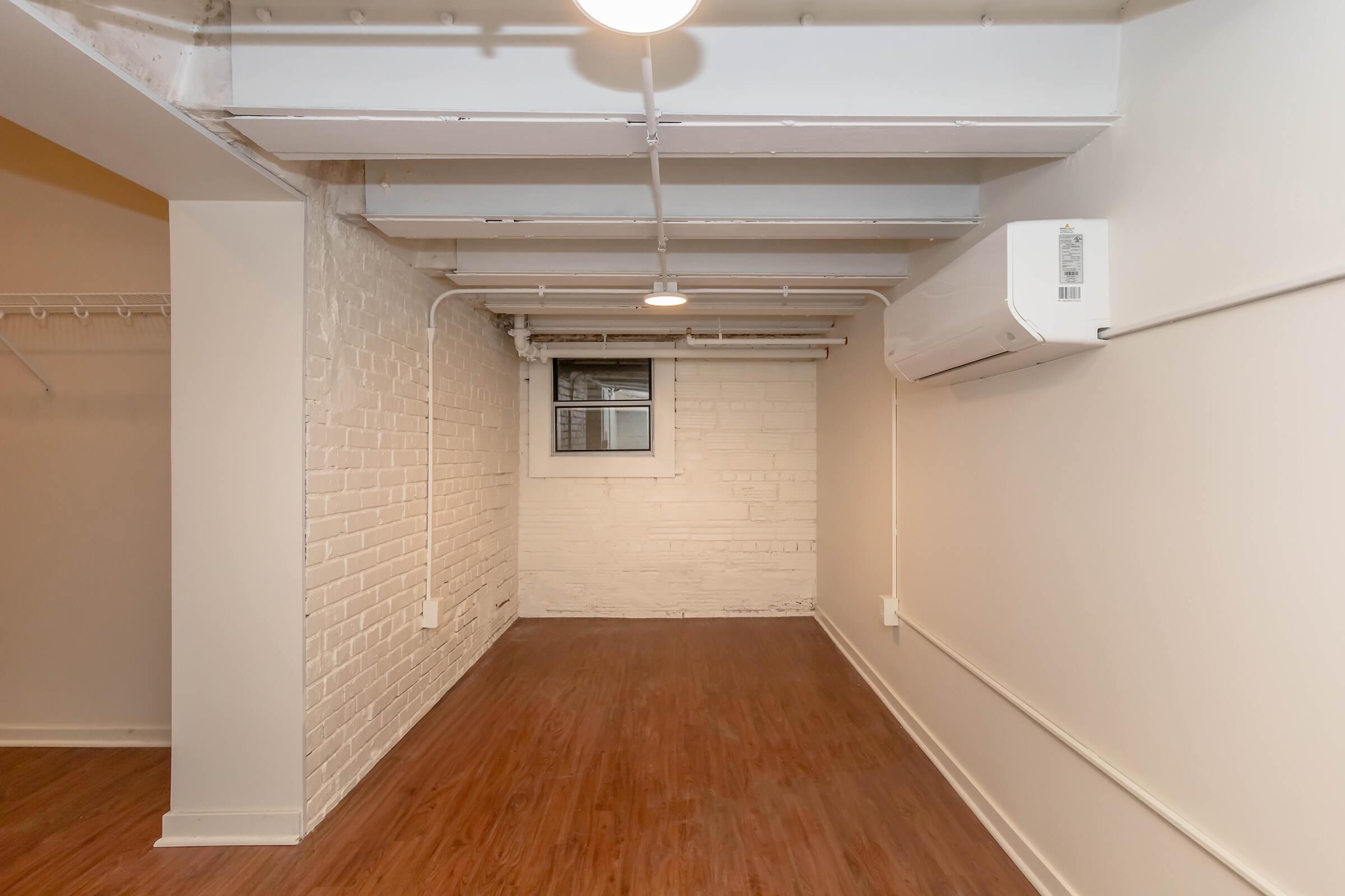 A bright, empty room with hardwood flooring and white brick walls. The space features exposed ceiling beams, a window with a view, and an air conditioning unit mounted on the wall. There's a small closet area visible on the left side of the image.