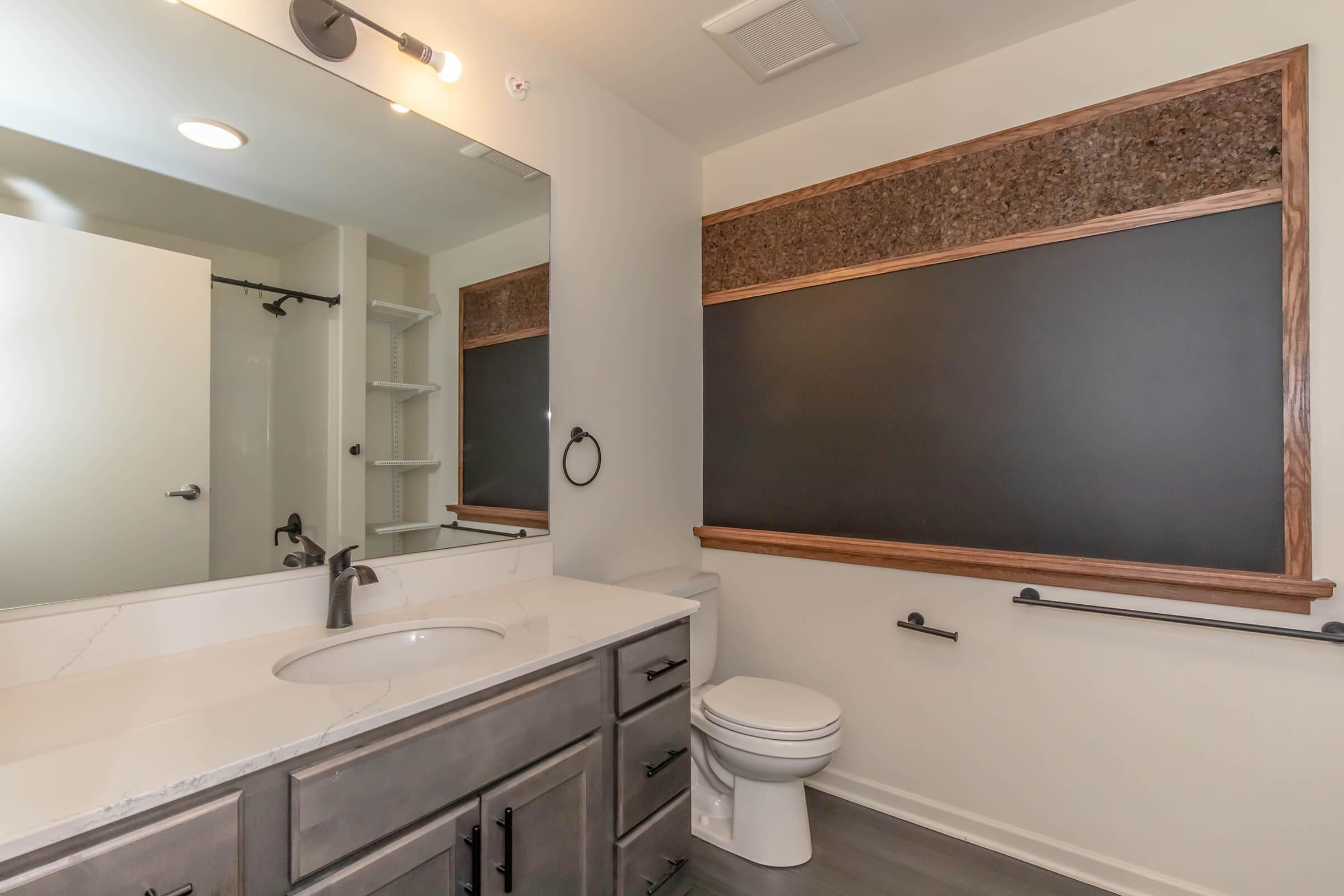A modern bathroom featuring a vanity with a marble countertop, a large mirror above, and a chalkboard panel framed in wood. The room includes a toilet and a shower area, with overhead lighting and a neutral color scheme. Shelving is visible to one side, adding to the functional design.
