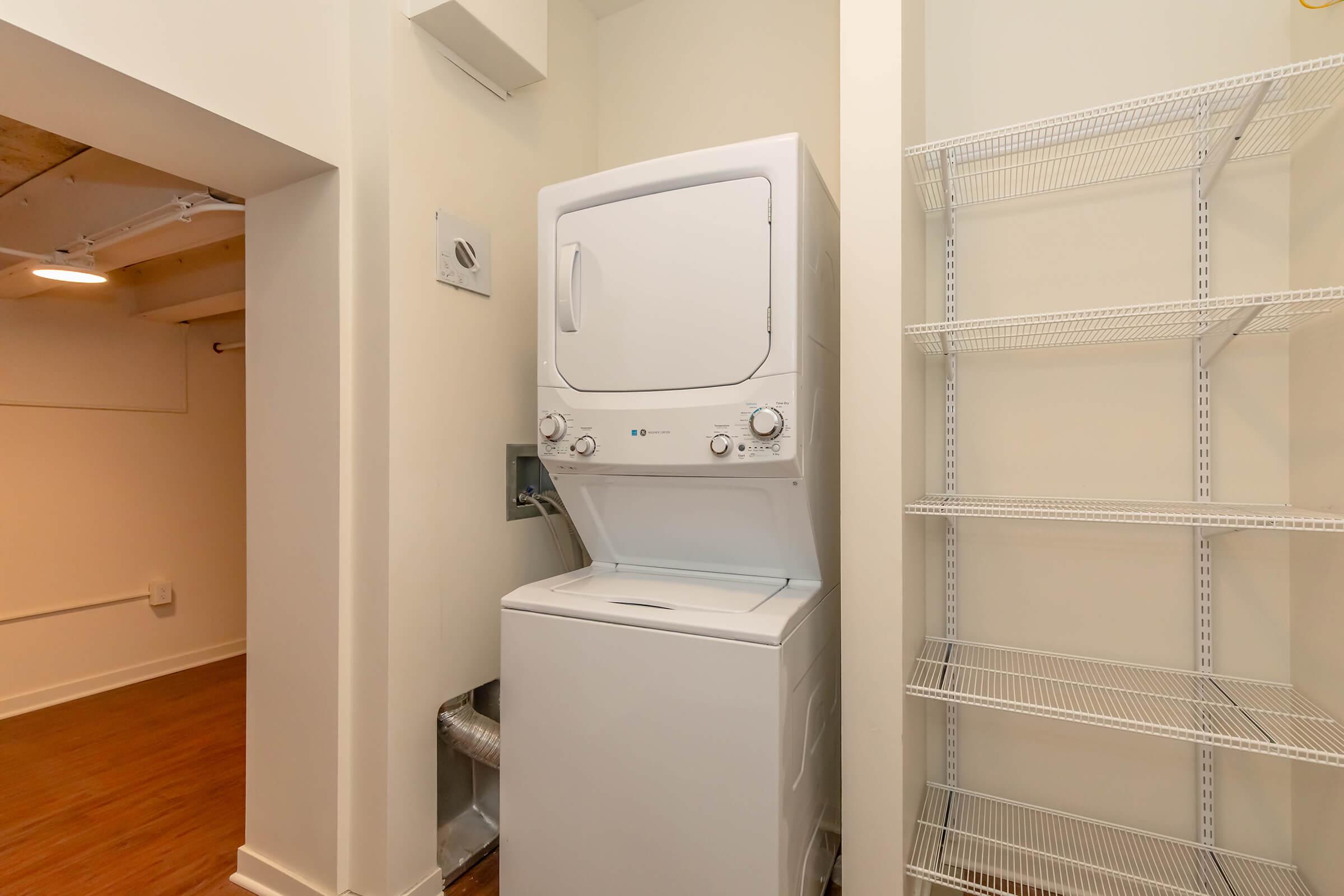 A compact laundry area featuring a stacked washer and dryer unit, positioned against a light-colored wall. Next to the appliances, there are metal wire shelves for storage. The floor is wooden, and the space is well-lit, creating a functional laundry nook.