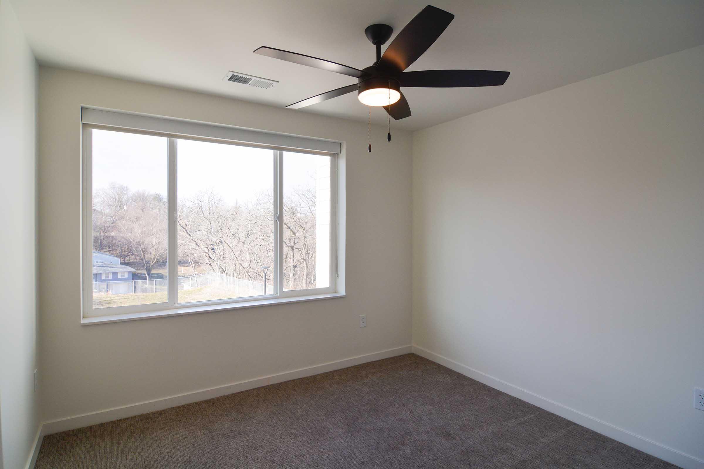 A bright, empty room with light beige carpet and a large window overlooking a tree-filled landscape. A modern ceiling fan with wooden blades hangs from the ceiling, adding a touch of style to the minimalist design. The walls are painted white, creating a spacious and airy feel.
