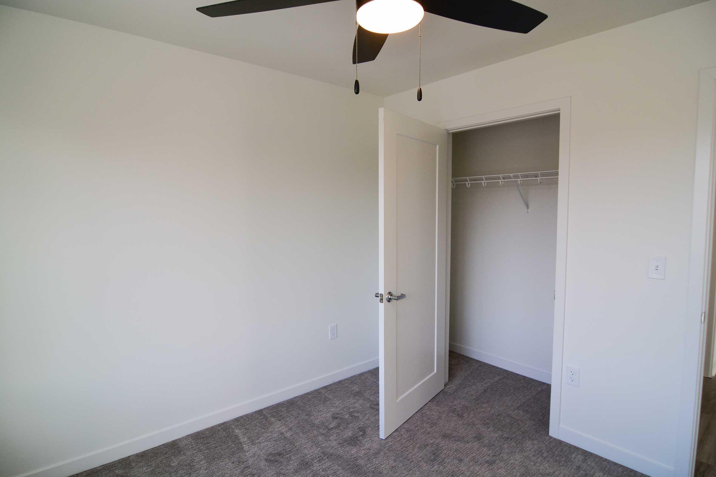 Empty room with light-colored walls and carpeted floor. A ceiling fan is visible, and there is an open closet with a single hanging rod. The door to the closet is on the right side, and the room has a clean, minimalistic design.