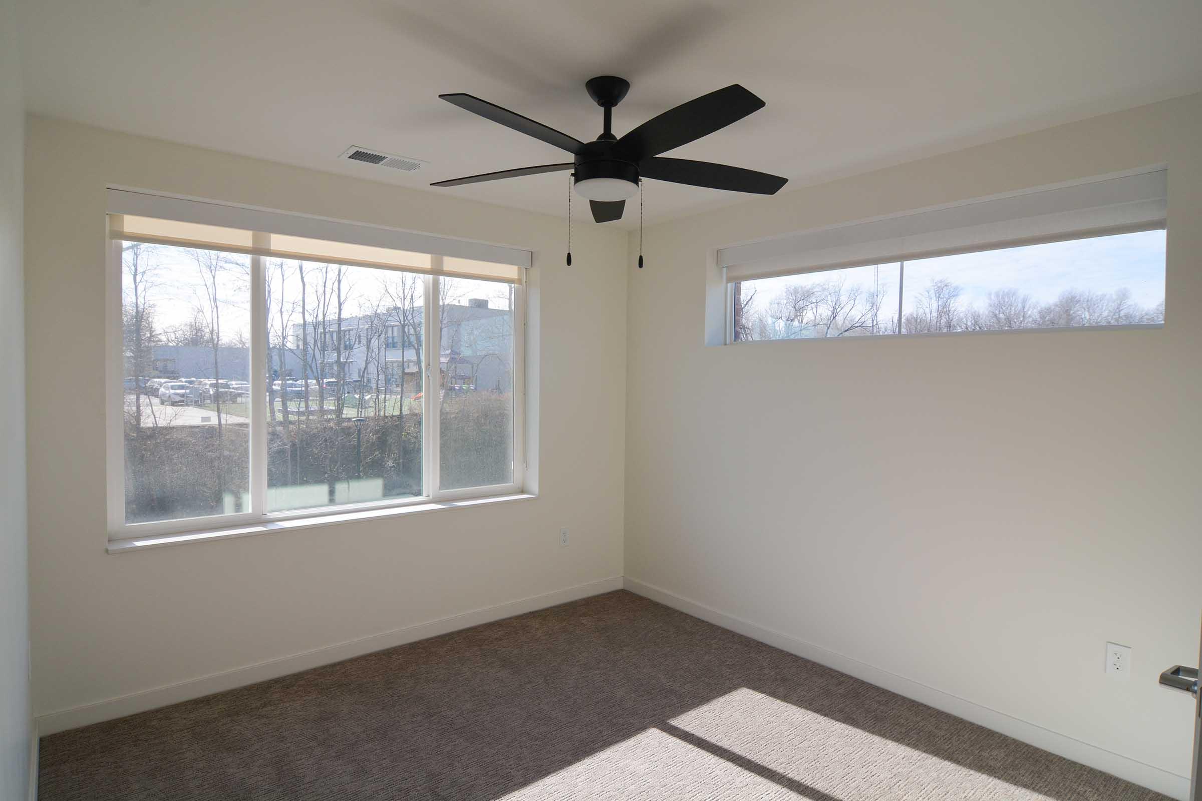 Empty room with beige carpet and light-colored walls. It features two large windows with white blinds, allowing natural light to enter. A black ceiling fan hangs from the ceiling, adding a modern touch to the space. The overall ambiance is bright and minimalist.