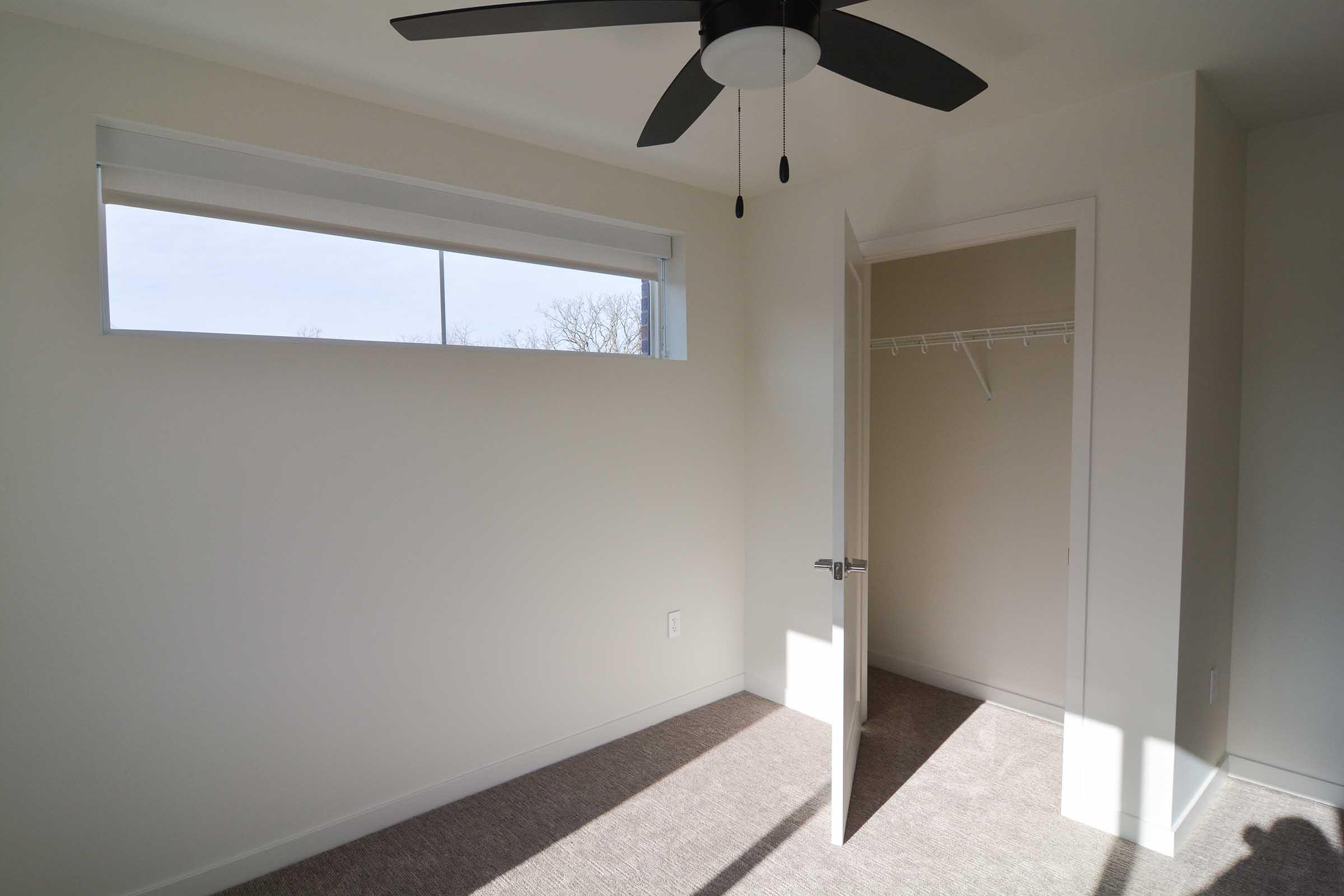 A bright, empty room featuring a ceiling fan, a large window with natural light streaming in, beige walls, and carpeted flooring. There's an open door leading to a closet with a hanging rack. The overall atmosphere is clean and minimalistic.