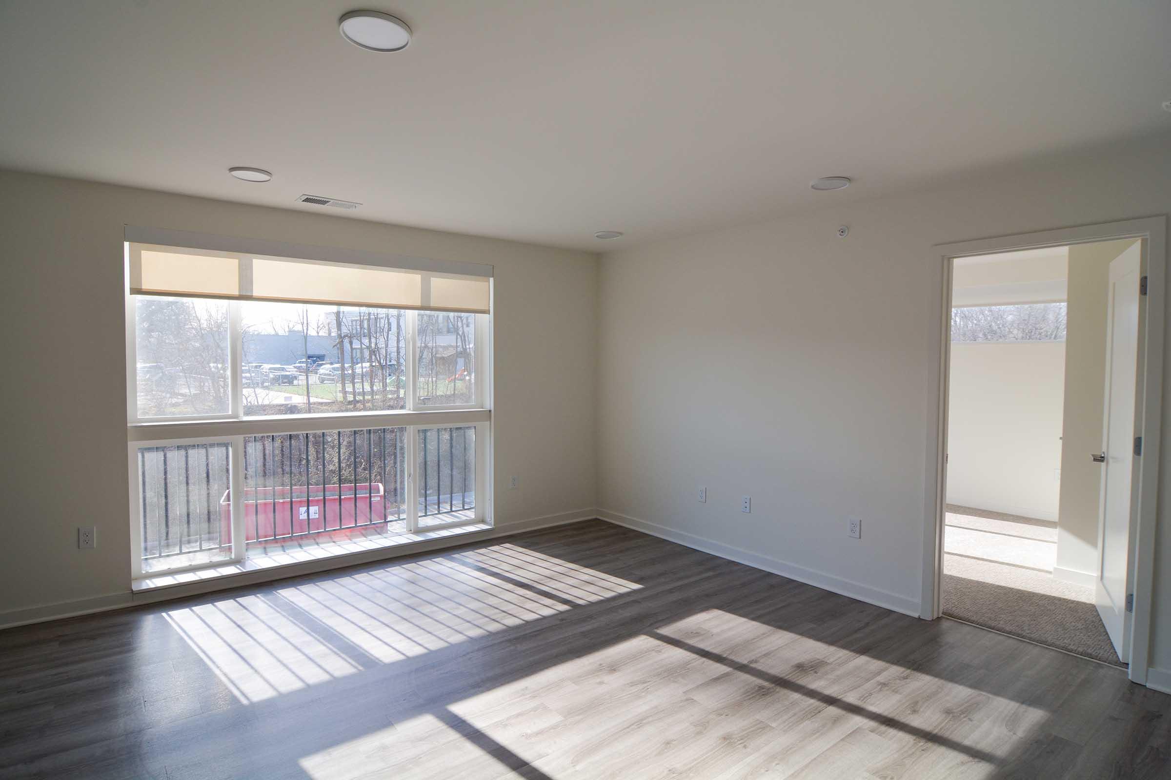 A bright, empty room featuring large windows with blinds, allowing natural light to fill the space. The floor is made of light brown laminate, and there is a door leading to another room. The walls are painted in a neutral color, creating a spacious and airy atmosphere.