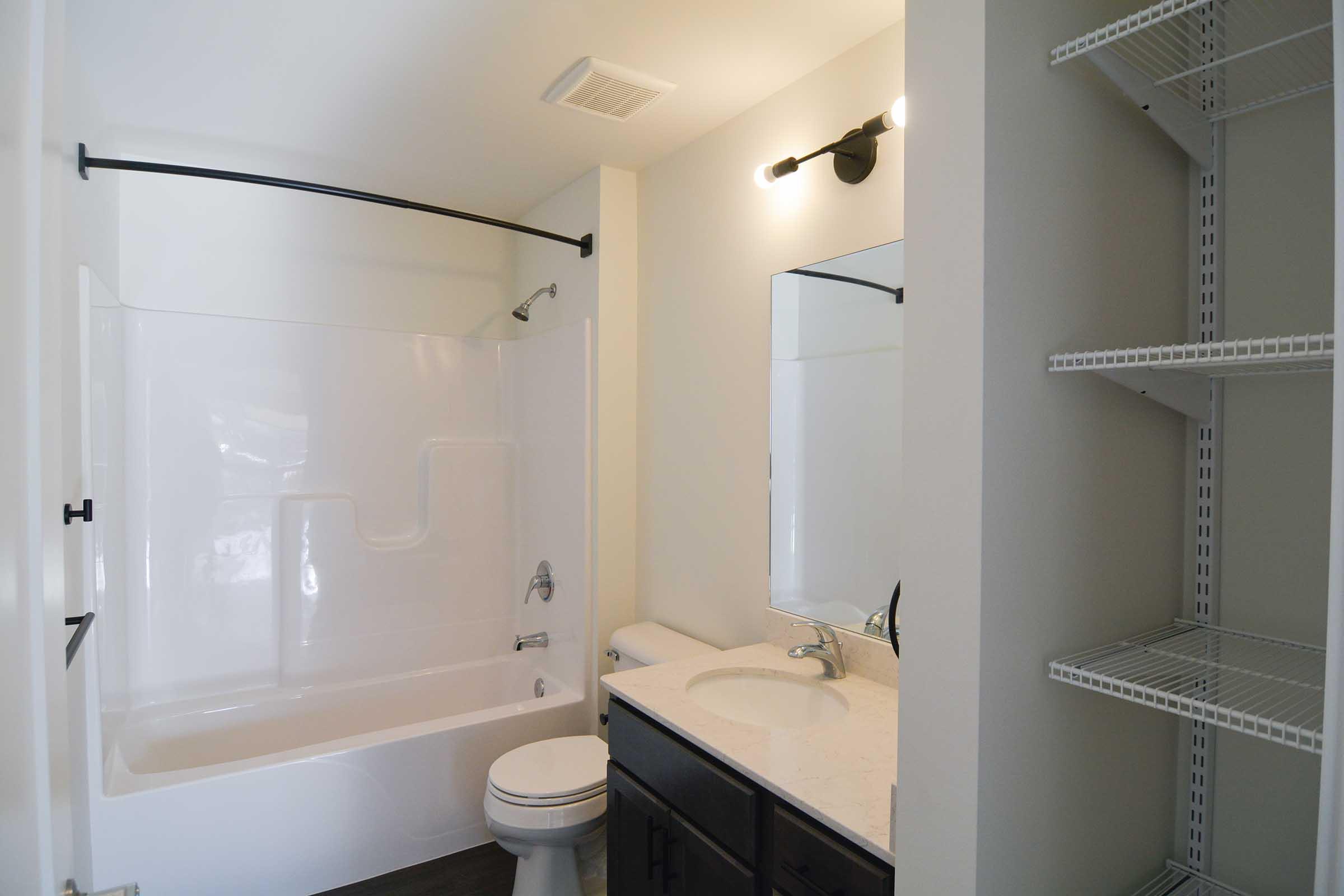 A modern bathroom featuring a bathtub with a shower, a toilet, a double sink vanity with a mirror, and white shelving. The walls are painted light colors, and there is ample natural light. The design is clean and minimalist.