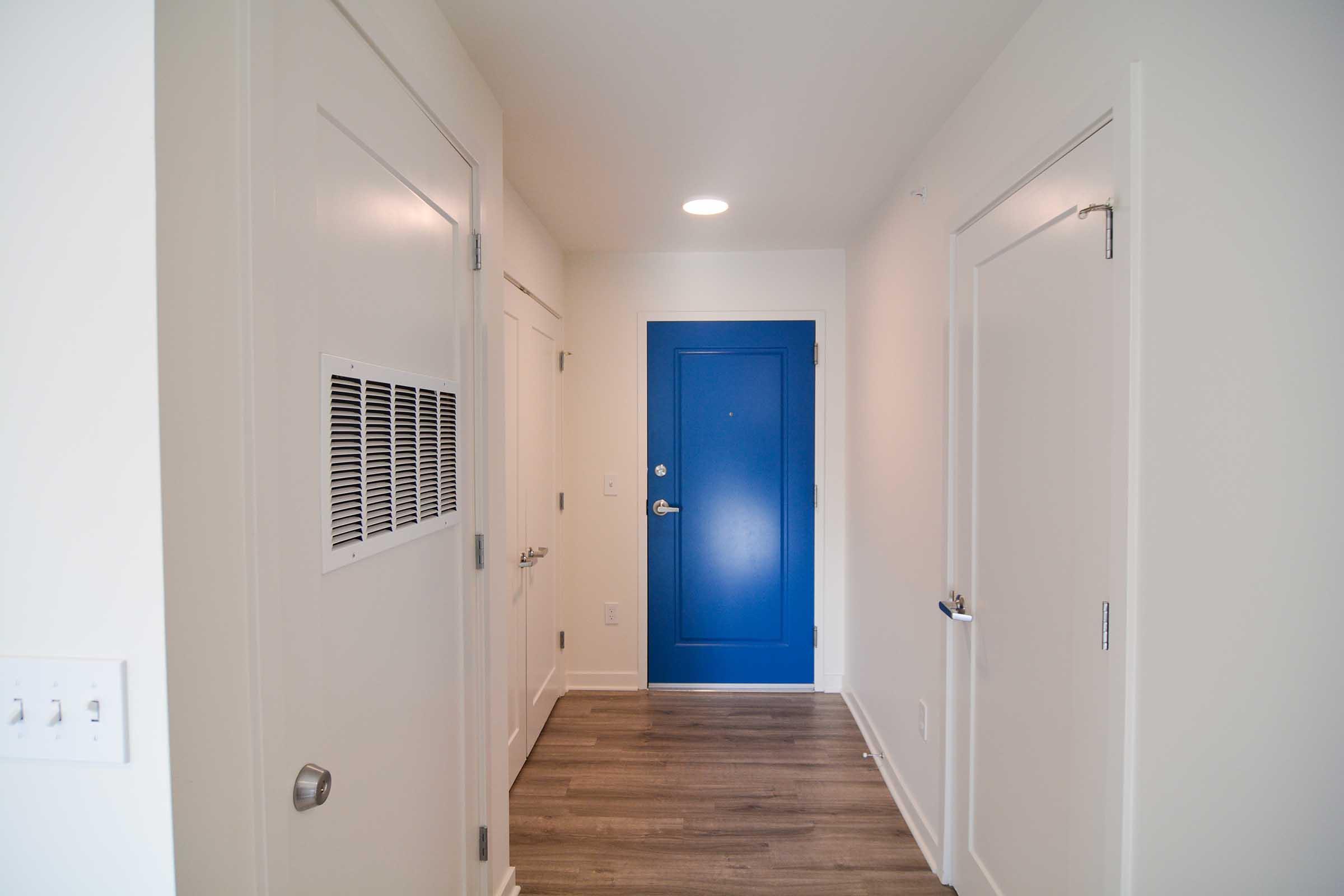 A well-lit hallway featuring light-colored walls and wooden flooring. To the left, there are two closed doors, and straight ahead is a bright blue front door. There is a ventilation grille on the wall, and the overall aesthetic is modern and inviting.