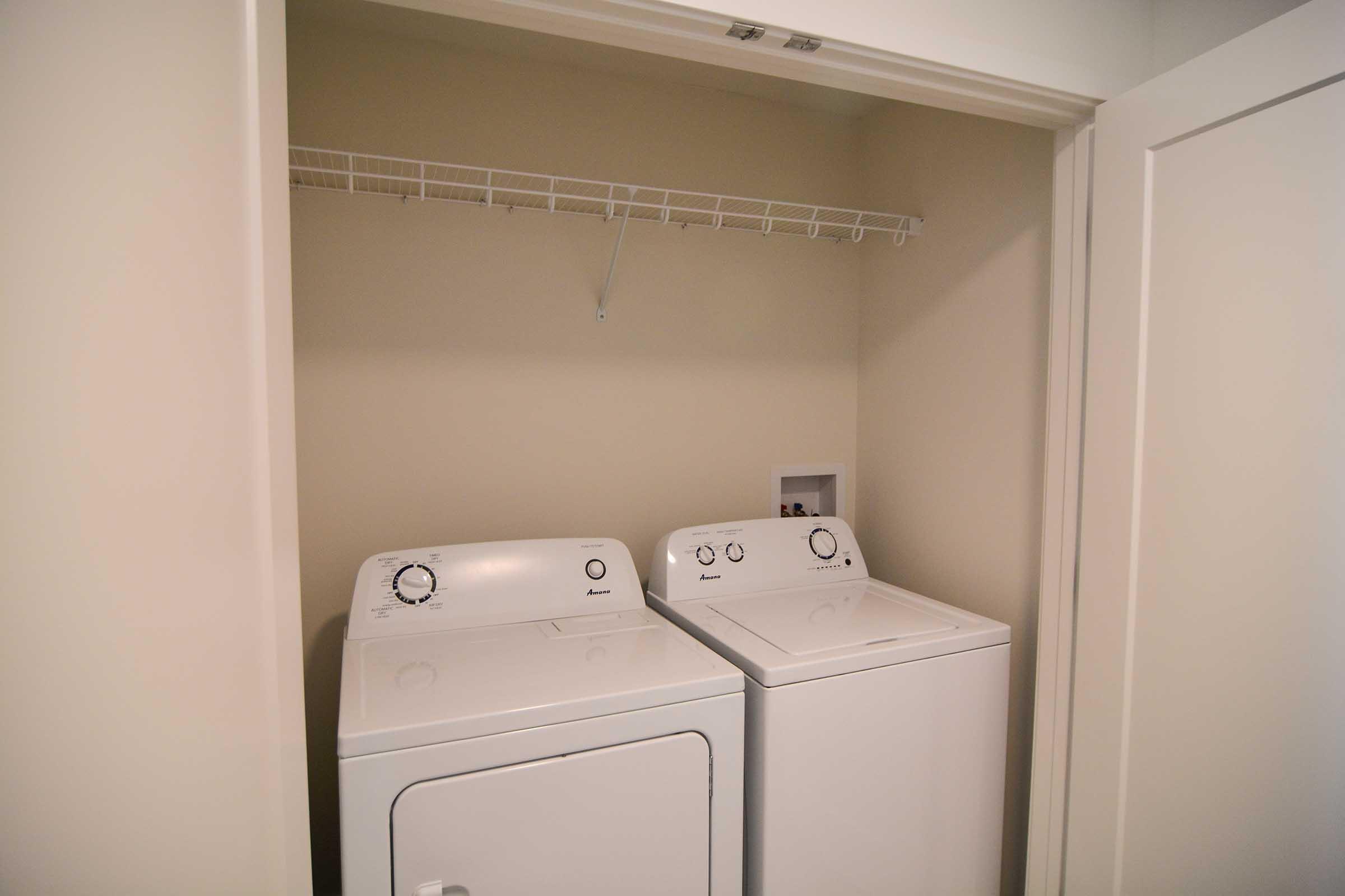 A compact laundry area featuring a stacked washer and dryer. The appliances are white and have visible control dials. Above them is a wire shelf for storage. The walls are painted a light color, and the closet has a sliding door that is partially open.