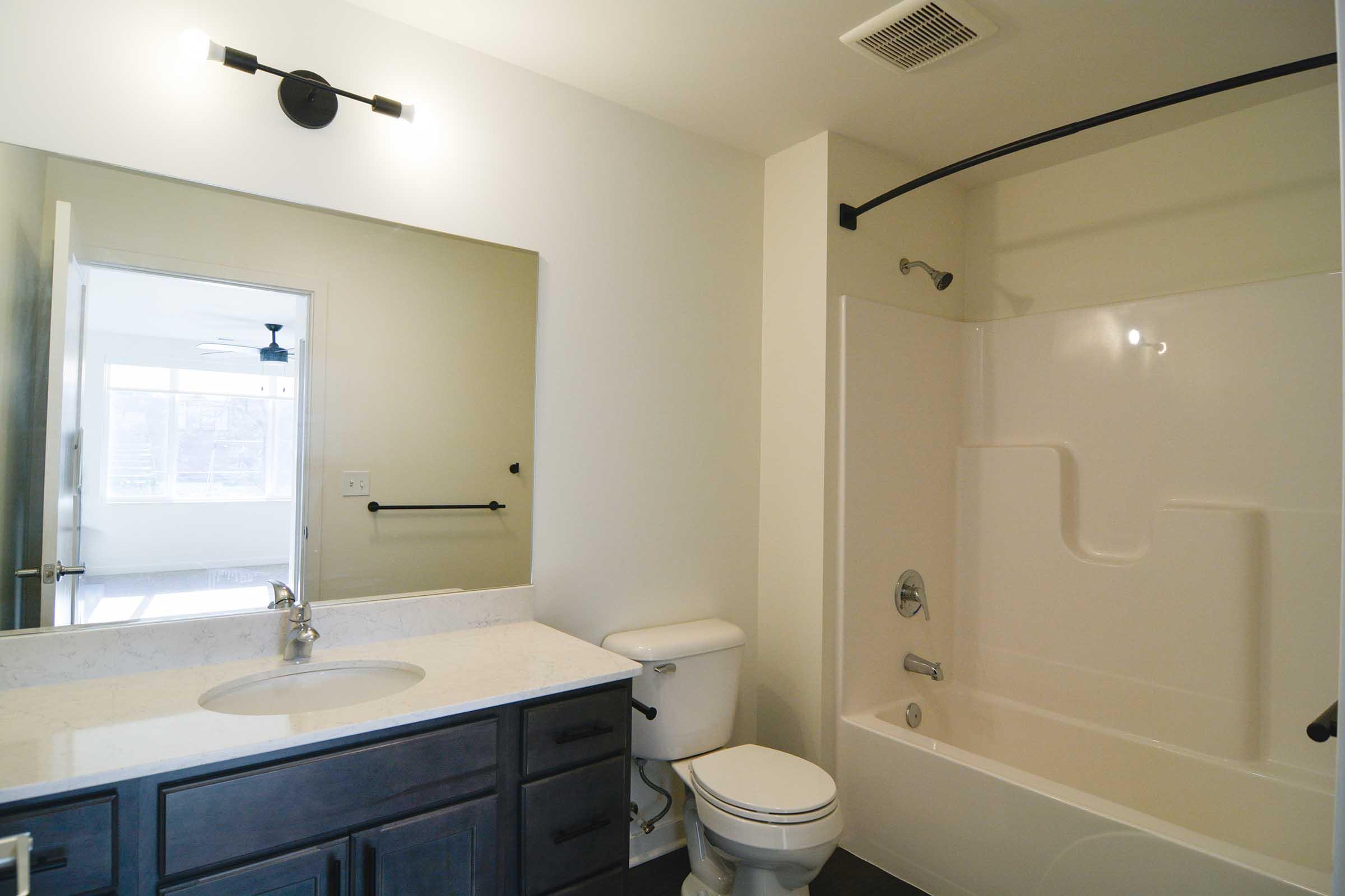 Modern bathroom featuring a white bathtub with a shower, a chrome faucet, a dark wood vanity with a marble countertop, and a large mirror. The space is well-lit with wall-mounted lights and has a door leading to another room. The flooring is dark, contrasting with the light walls.