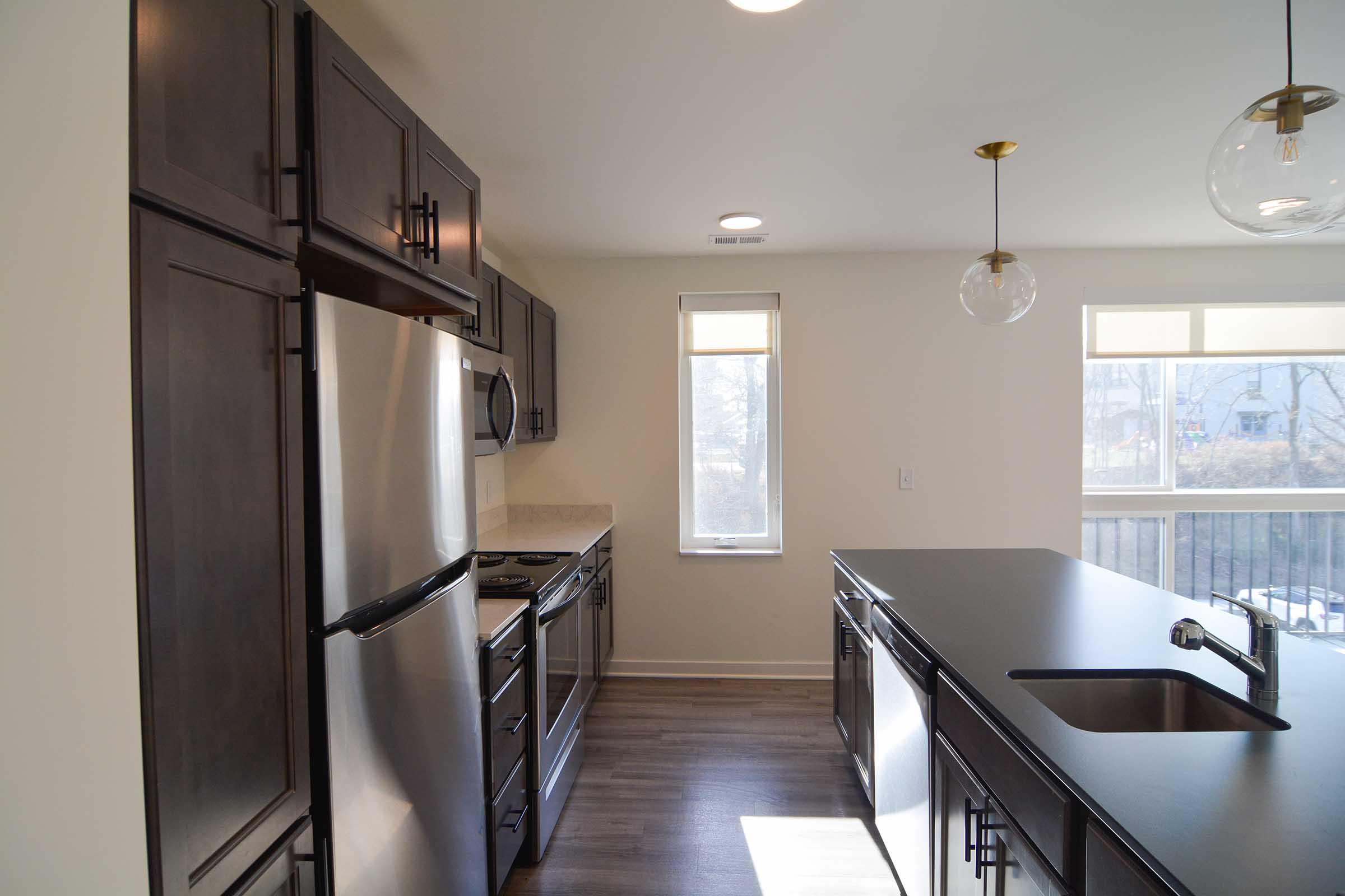Modern kitchen featuring dark wood cabinetry, stainless steel appliances including a refrigerator and stove, a central island with a sink, and pendant lighting. Large window providing natural light and a view of the outside. Clean, contemporary design with a neutral color palette.