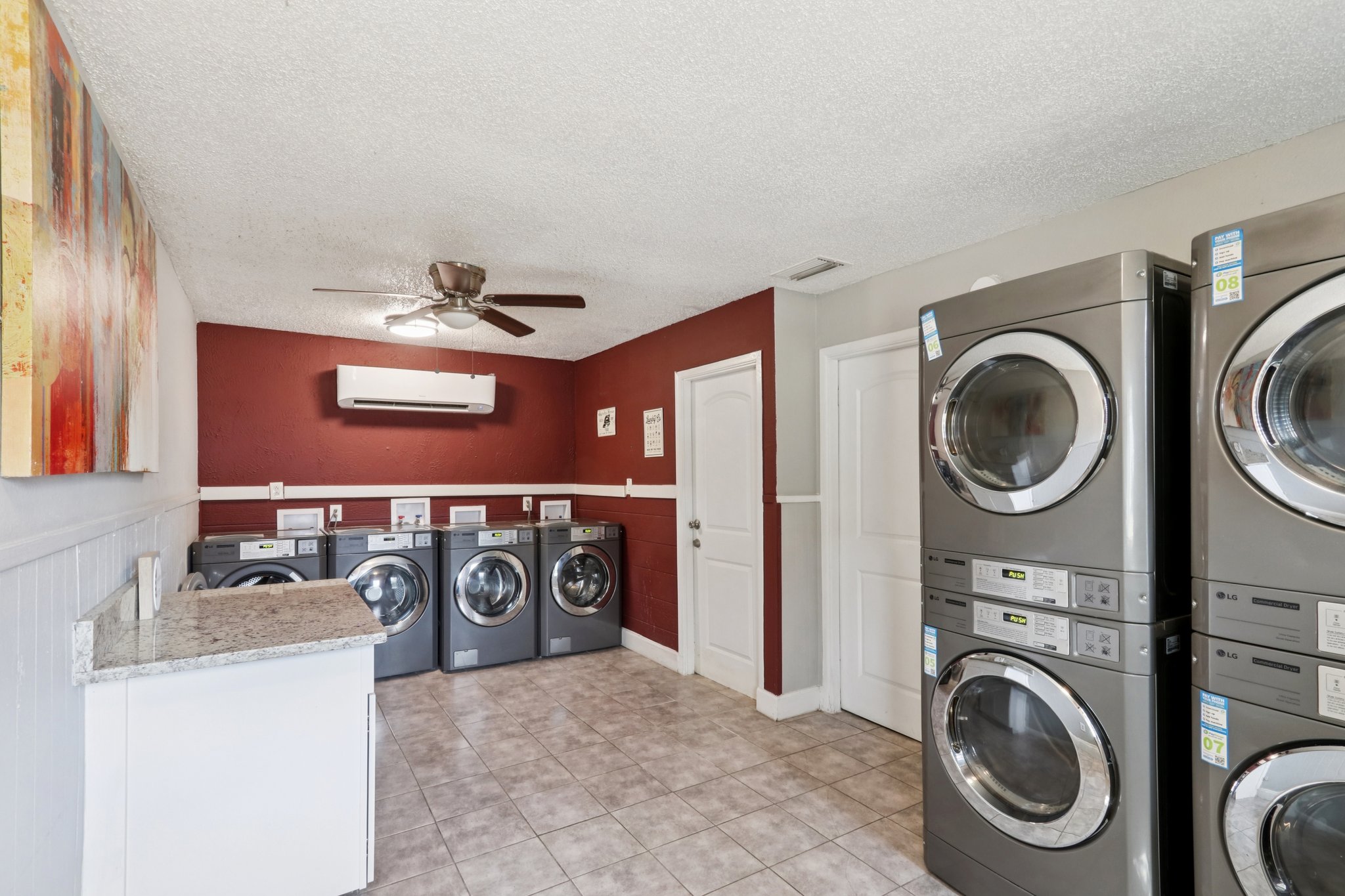Interior view of a laundry room featuring three stacked washer and dryer units in metallic finish, a white countertop, and a wall-mounted air conditioning unit. The walls are painted in a deep red color, and there is a ceiling fan for ventilation. A door leads to another area, and decorative artwork is visible on one wall.