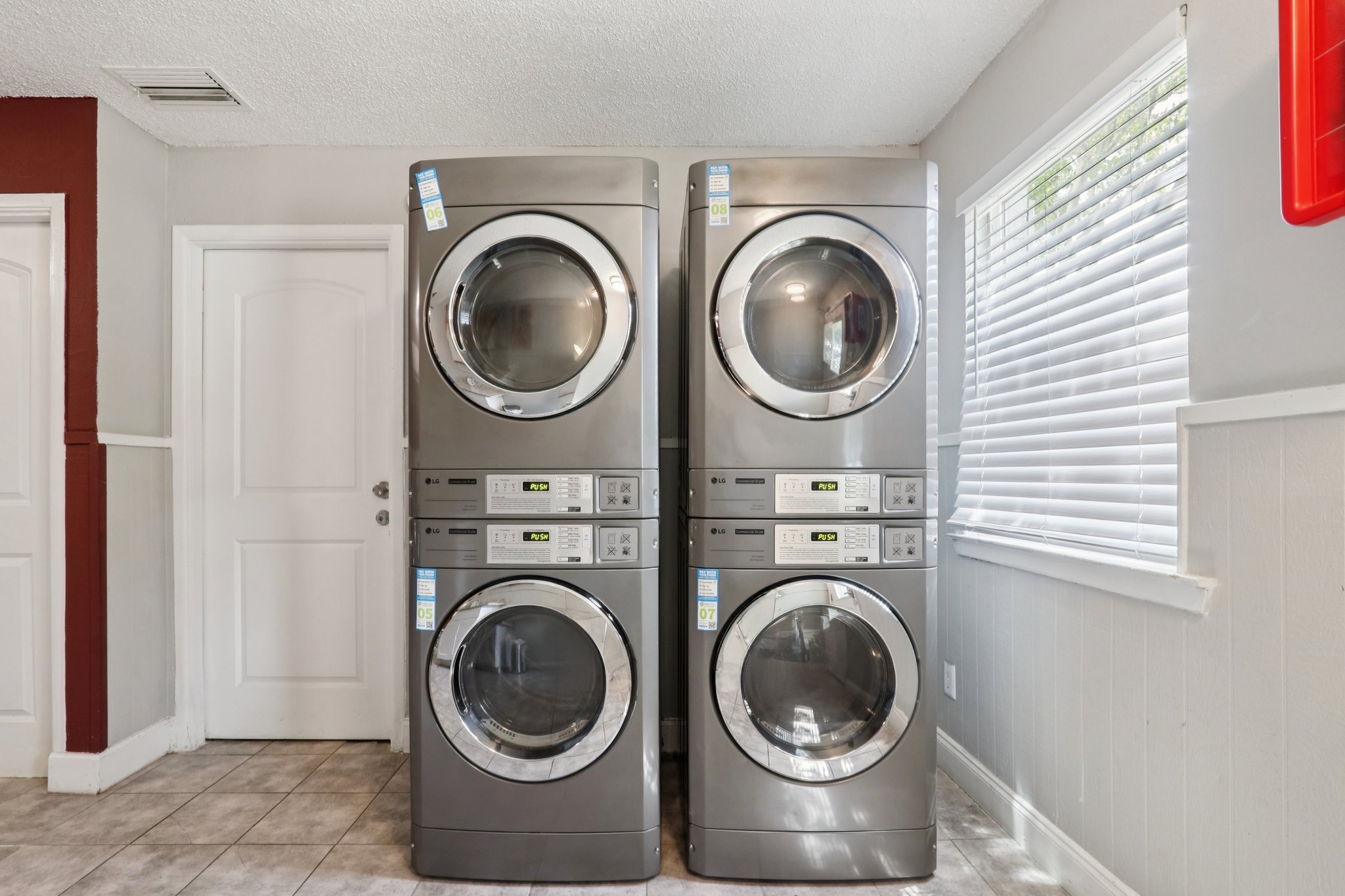 Two stacked gray washer and dryer units in a laundry room, positioned against a light gray wall. There is a white door on the left and a window with blinds allowing natural light in. The flooring is tiled in neutral colors, creating a functional and efficient laundry space.