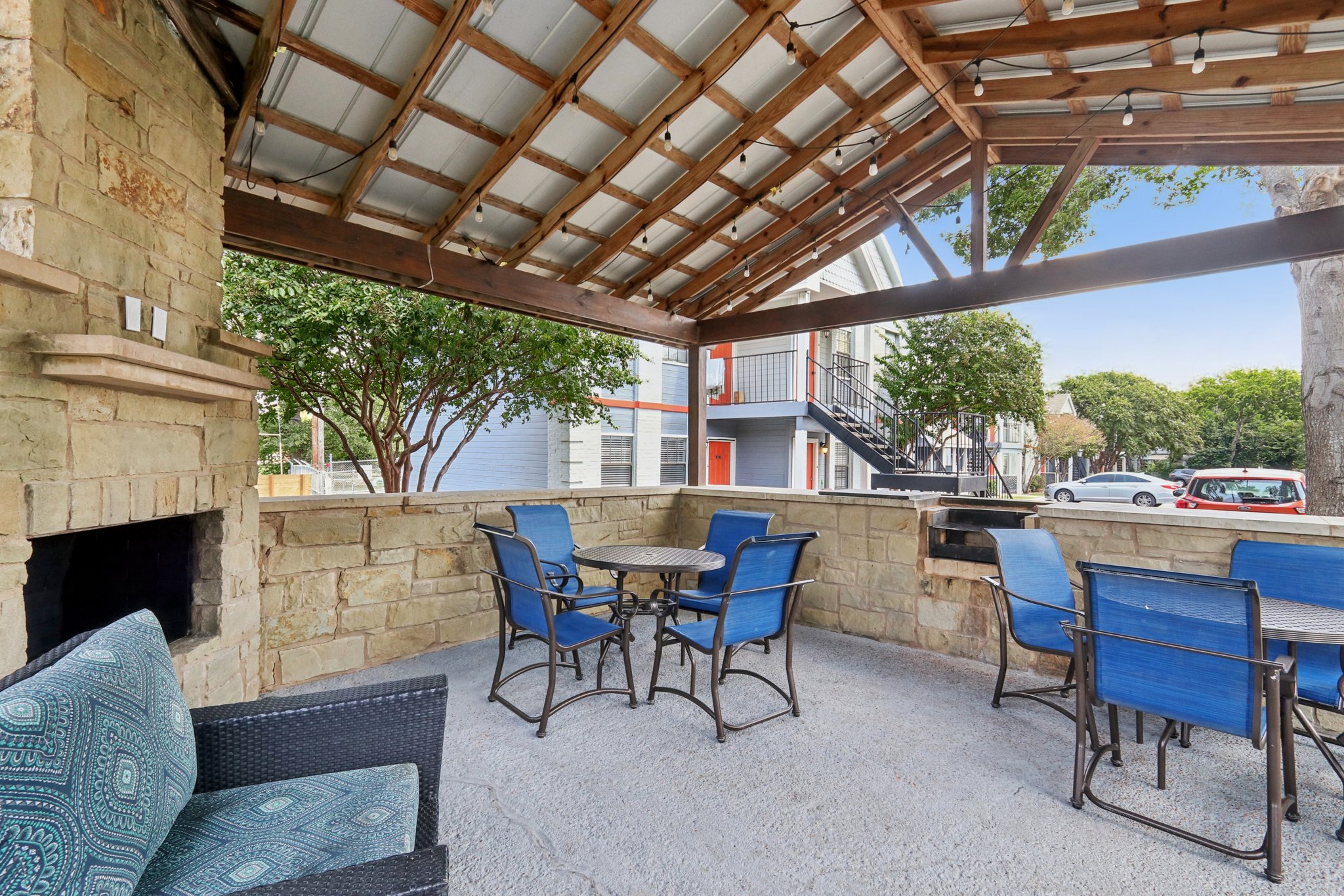 Outdoor covered patio area featuring a stone fireplace, a round table with blue chairs, and a textured rug. Surrounding trees provide shade, and a view of a building with balconies and red doors is visible in the background.