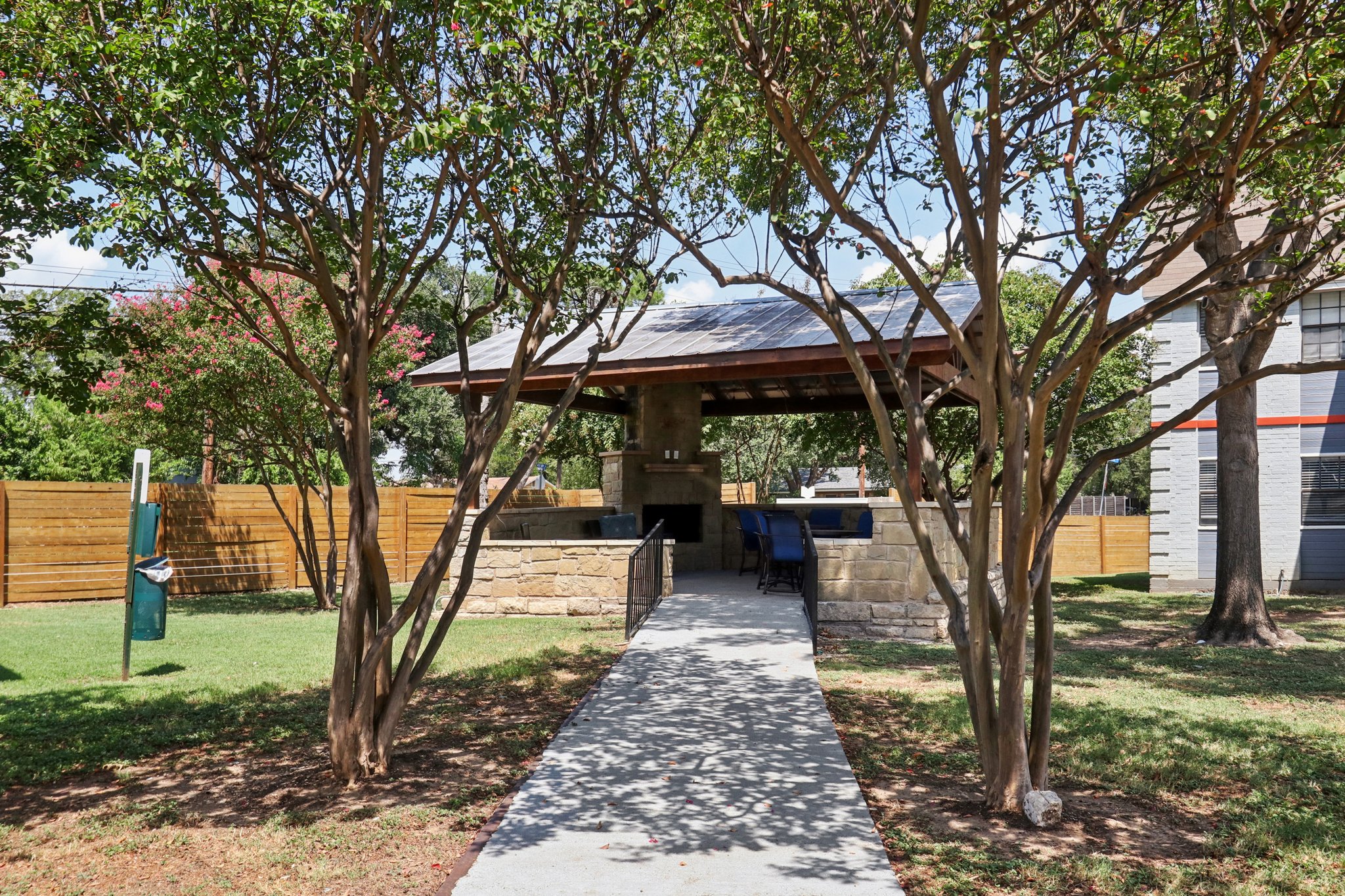 A shaded outdoor pavilion surrounded by trees and green grass. The pavilion features a stone structure with seating and an outdoor grill. A concrete pathway leads through the trees towards the pavilion. In the background, there are wooden fences and a building visible.
