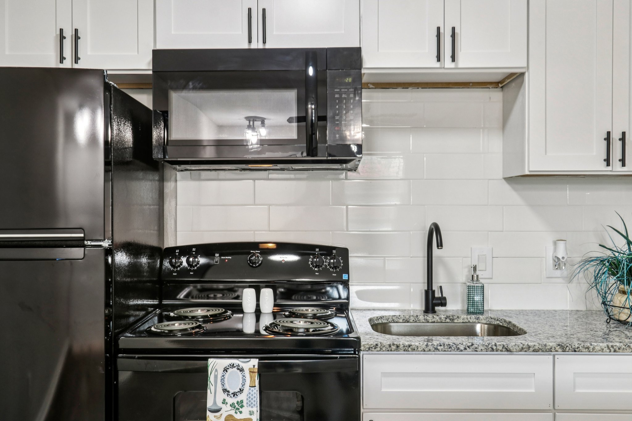 A modern kitchen featuring a black refrigerator, black stove, and a microwave mounted above. The countertop is granite with a stainless steel sink and a black faucet. White cabinets and tiled backsplash provide a bright, clean look, while a decorative plant adds a touch of color.