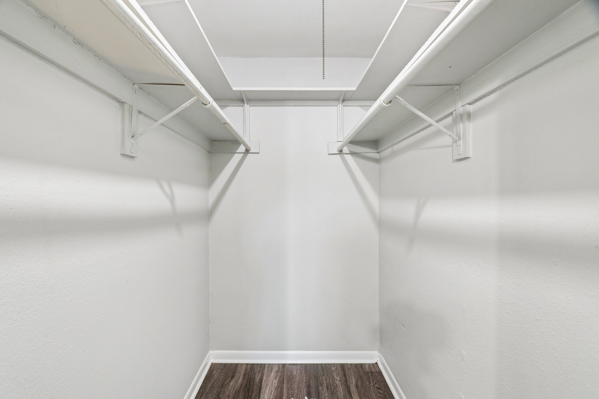 Empty closet with white walls and a wooden floor. The ceiling features two horizontal clothing rods for hanging items. There are no shelves or other furnishings in the space.