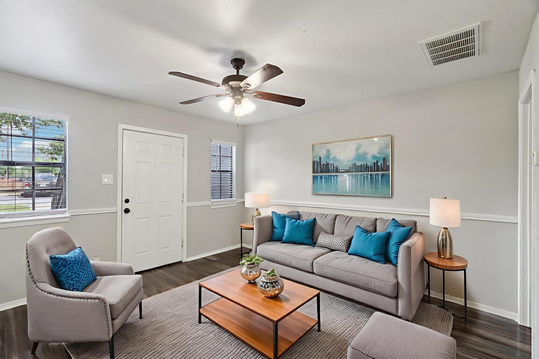 A cozy living room featuring a gray sofa with blue accent pillows, a wooden coffee table, and a stylish lamp. A ceiling fan provides illumination, while large windows allow natural light to fill the space. Artwork depicting a city skyline adds an artistic touch to the wall.