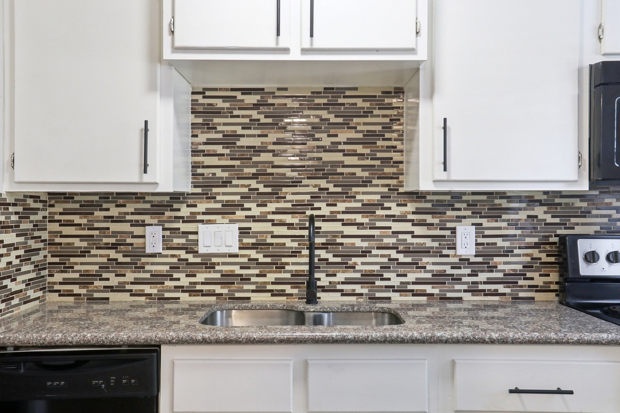 Modern kitchen featuring a stainless steel sink with a sleek black faucet, set against a decorative mosaic tile backsplash in earthy tones. The cabinetry above is white with minimalistic black handles, and there’s a black stove to the right, adding to the contemporary feel of the space.