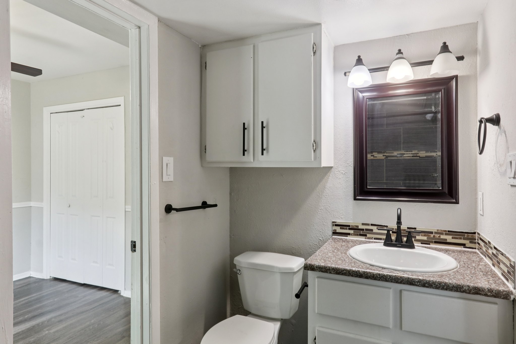 A modern bathroom featuring a white toilet, a granite countertop with a sink, and a large mirror. The walls are painted a light color, and there is a cabinet above the sink. Lighting fixtures with three bulbs hang above the mirror, and a towel bar is mounted on the wall beside the sink.