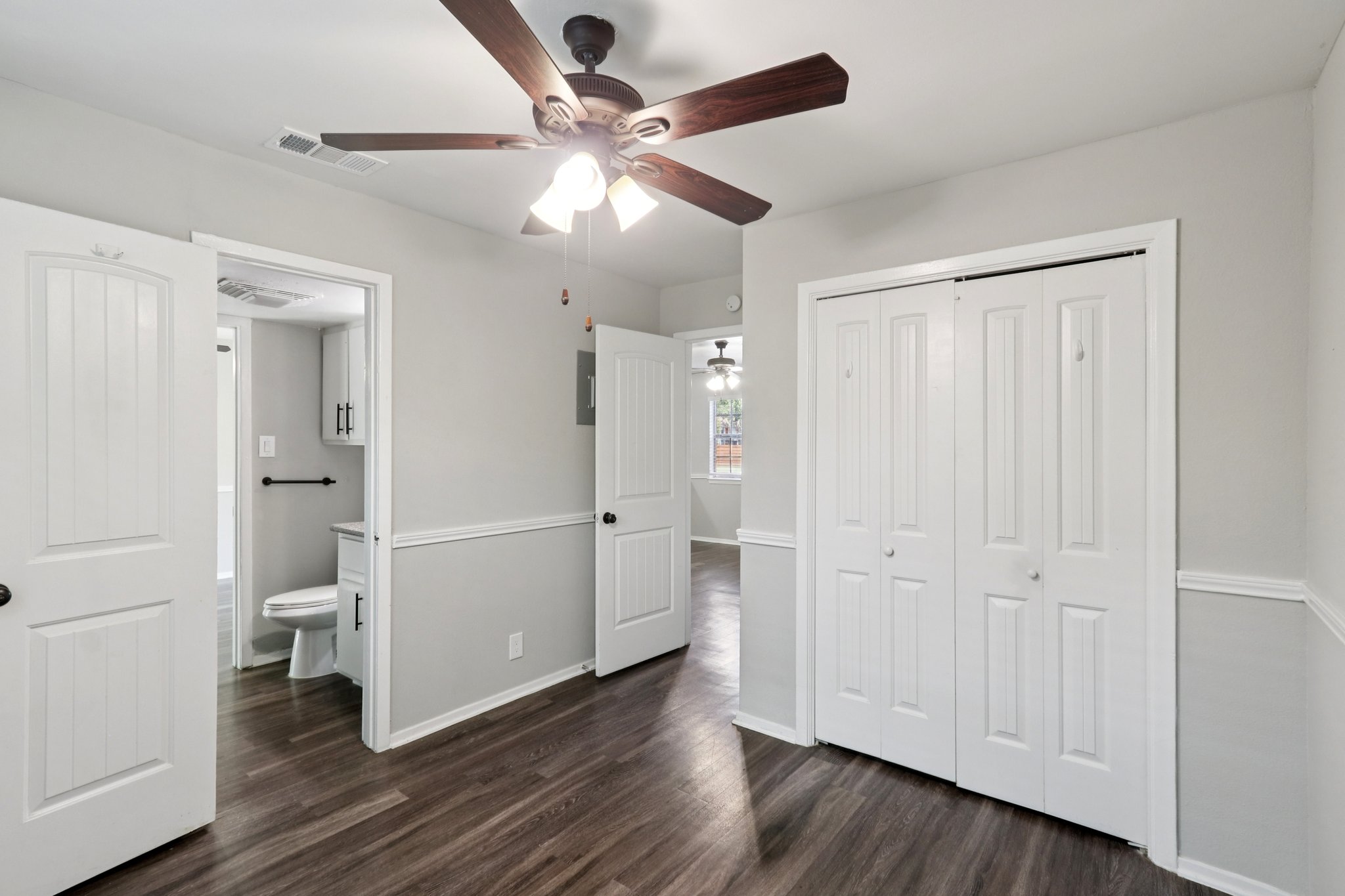 Interior view of a bright, modern room featuring a ceiling fan, two doors leading to closets or other spaces, and a small bathroom entrance visible in the background. The walls are painted in a light color, and the flooring is dark wood, creating a contrast in the space.