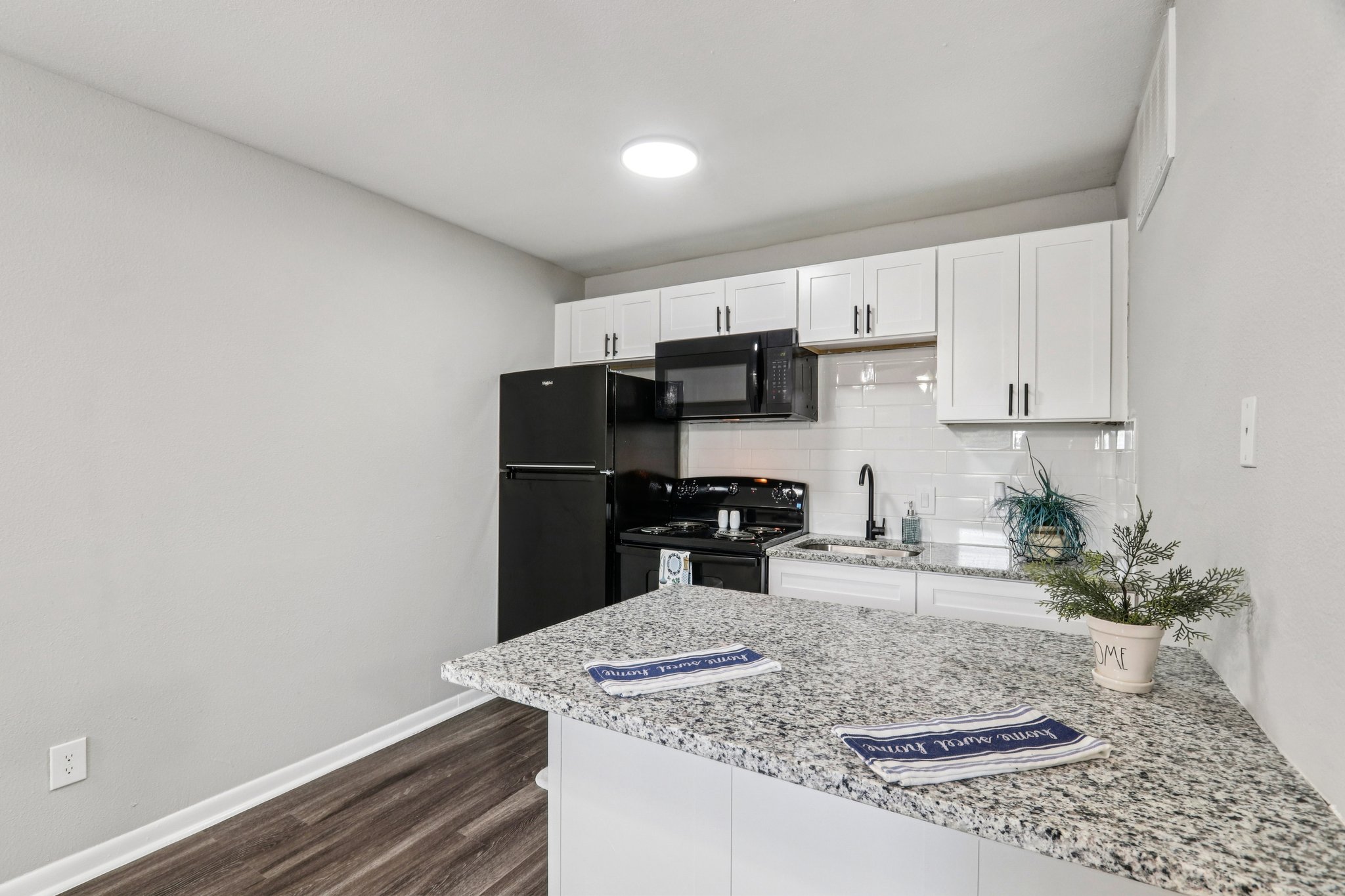 Modern kitchen featuring white cabinetry, a black refrigerator, and an oven with a microwave above. The countertops are made of gray speckled granite. A small plant is on the counter, and there are two brochures laid out. The flooring is a dark wood laminate, and the walls are painted light gray.