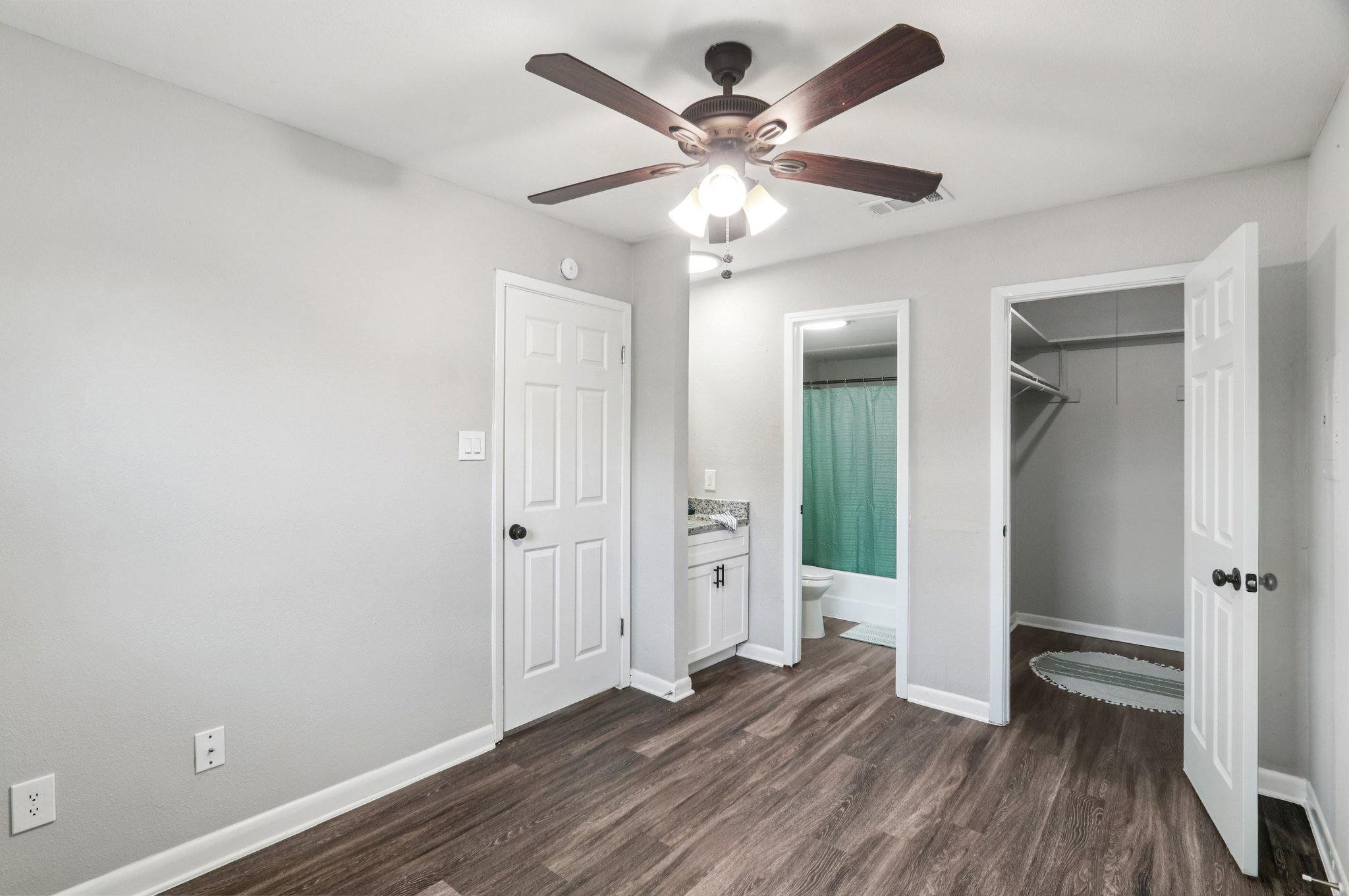 A well-lit bedroom featuring light gray walls and a ceiling fan. The room has a closet with open doors and a bathroom visible in the background. The flooring is brown laminate, and there is a round rug near the closet. The space appears clean and modern, ideal for relaxation.