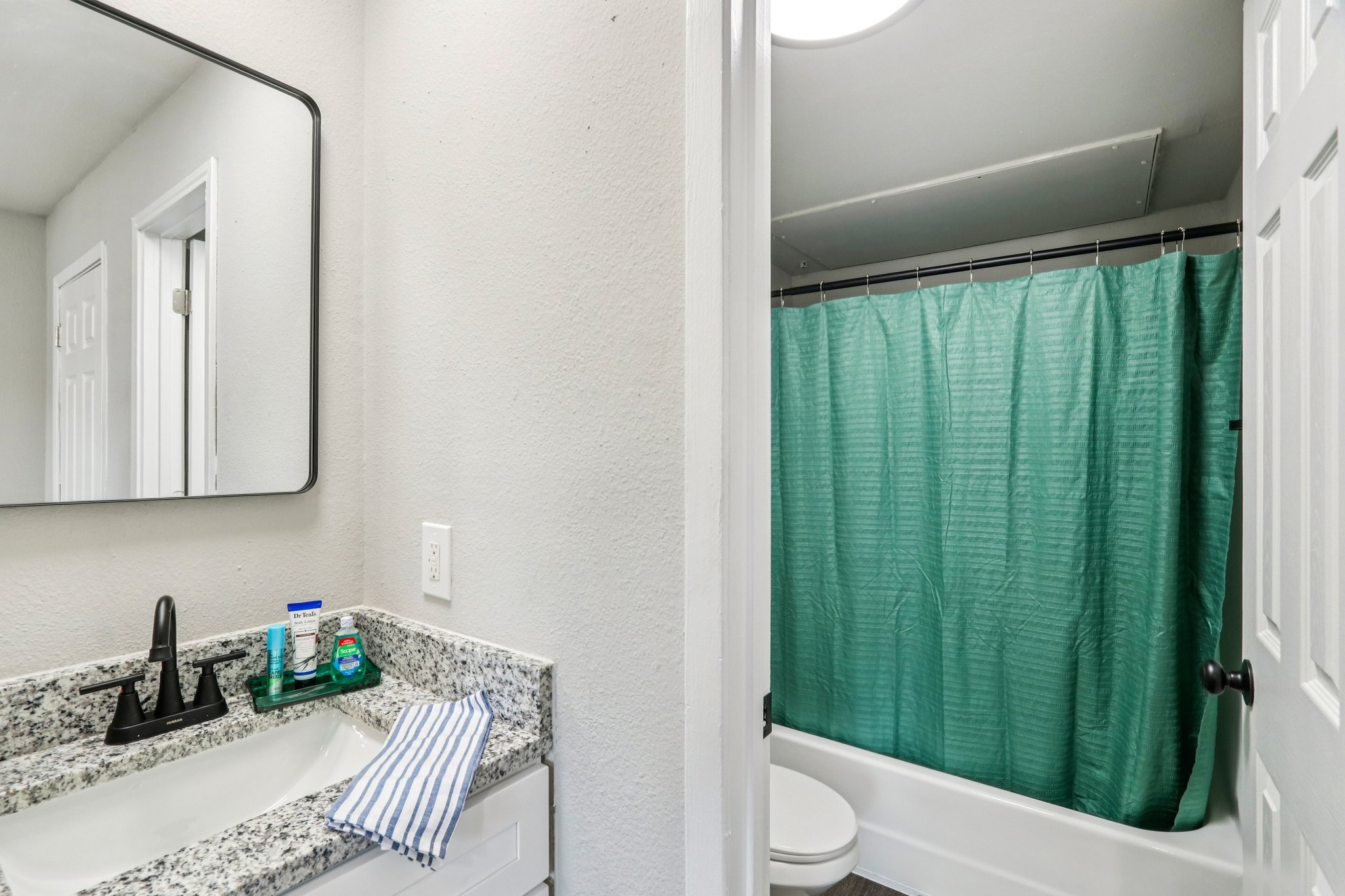 A clean bathroom featuring a granite countertop with a sink, a modern faucet, and a mirror above. On the countertop, there is a striped towel and toiletries. To the right, a shower area with a green curtain and a white bathtub is visible. The walls are painted in a light color, giving a bright and spacious appearance.
