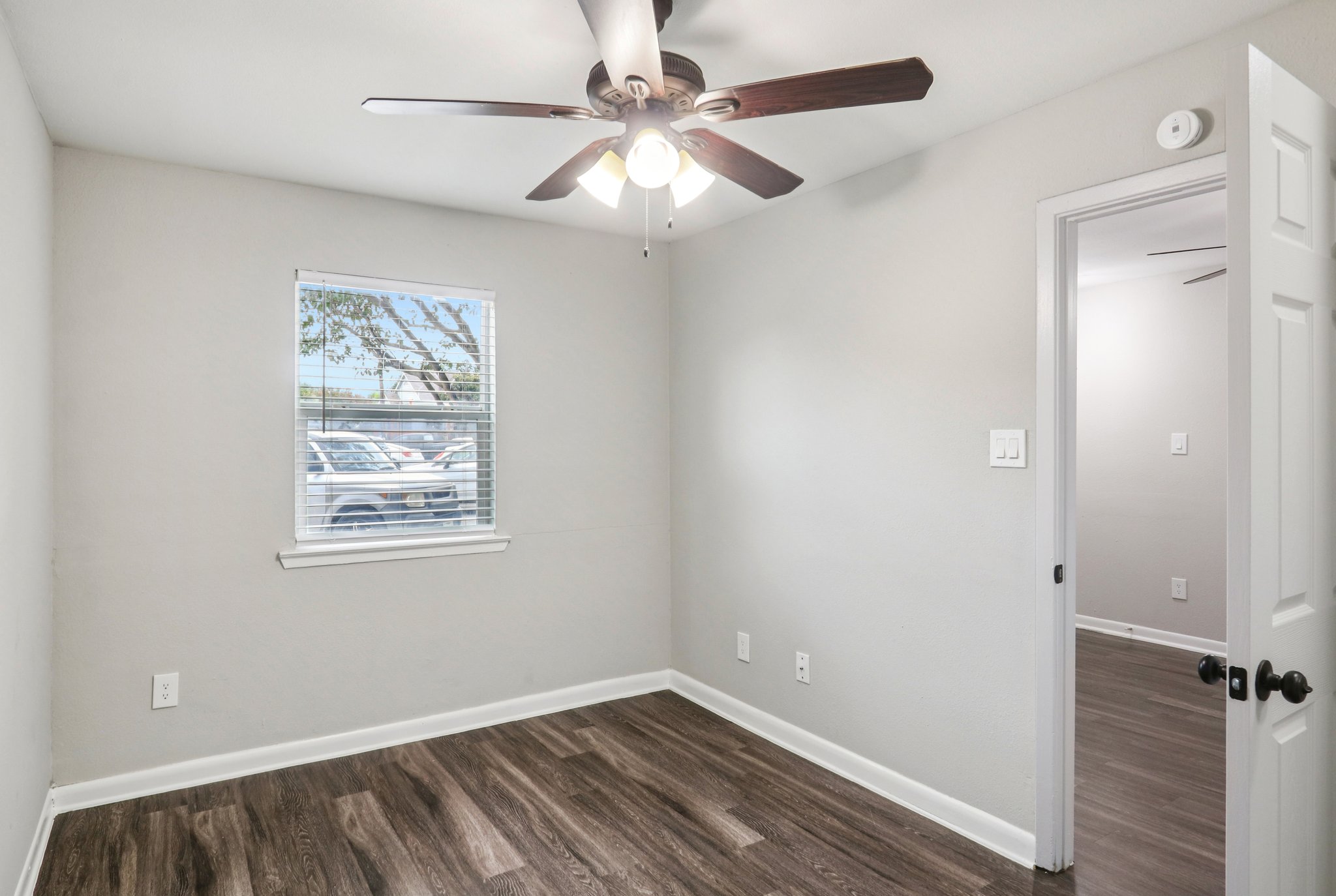 A small, minimally furnished room with light gray walls and brown laminate flooring. A ceiling fan with light hangs from the ceiling. There’s a window with blinds allowing natural light, and a door on the right leads to another room. The space is tidy and bright, with a few visible cars outside through the window.