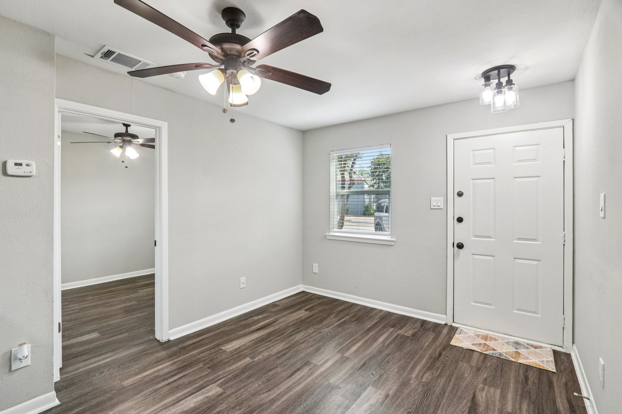 A bright, freshly painted room with light gray walls and dark laminate flooring. There are two ceiling fans with light fixtures, and a front door with a window beside it. Natural light comes through a window, illuminating the space, creating a clean and inviting atmosphere.