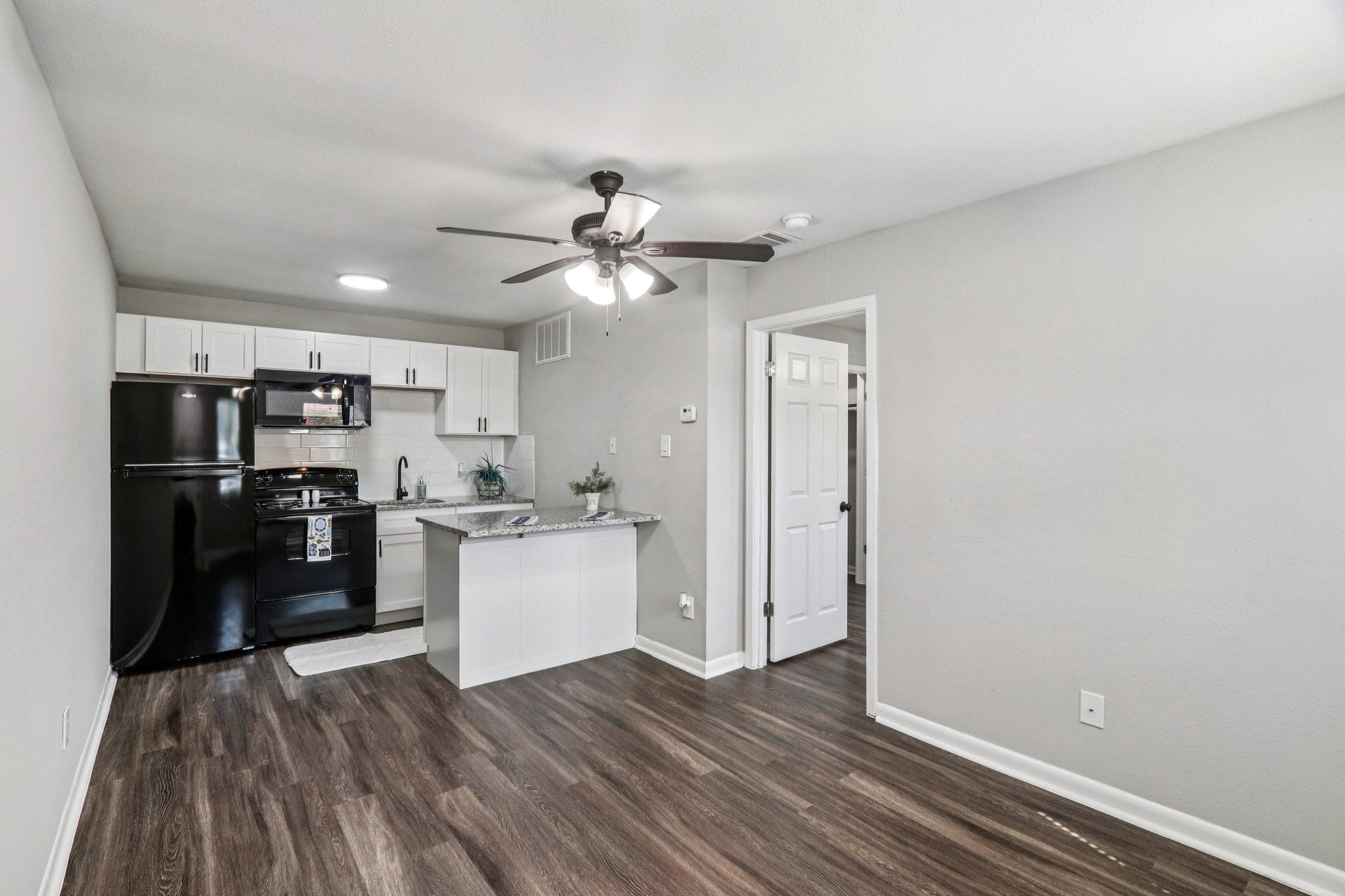 A modern, compact kitchen featuring black appliances, white cabinets, and a granite countertop. The space also includes a ceiling fan, wood-look flooring, and a doorway leading to another room. Natural light enters through a window, creating a bright and inviting atmosphere.