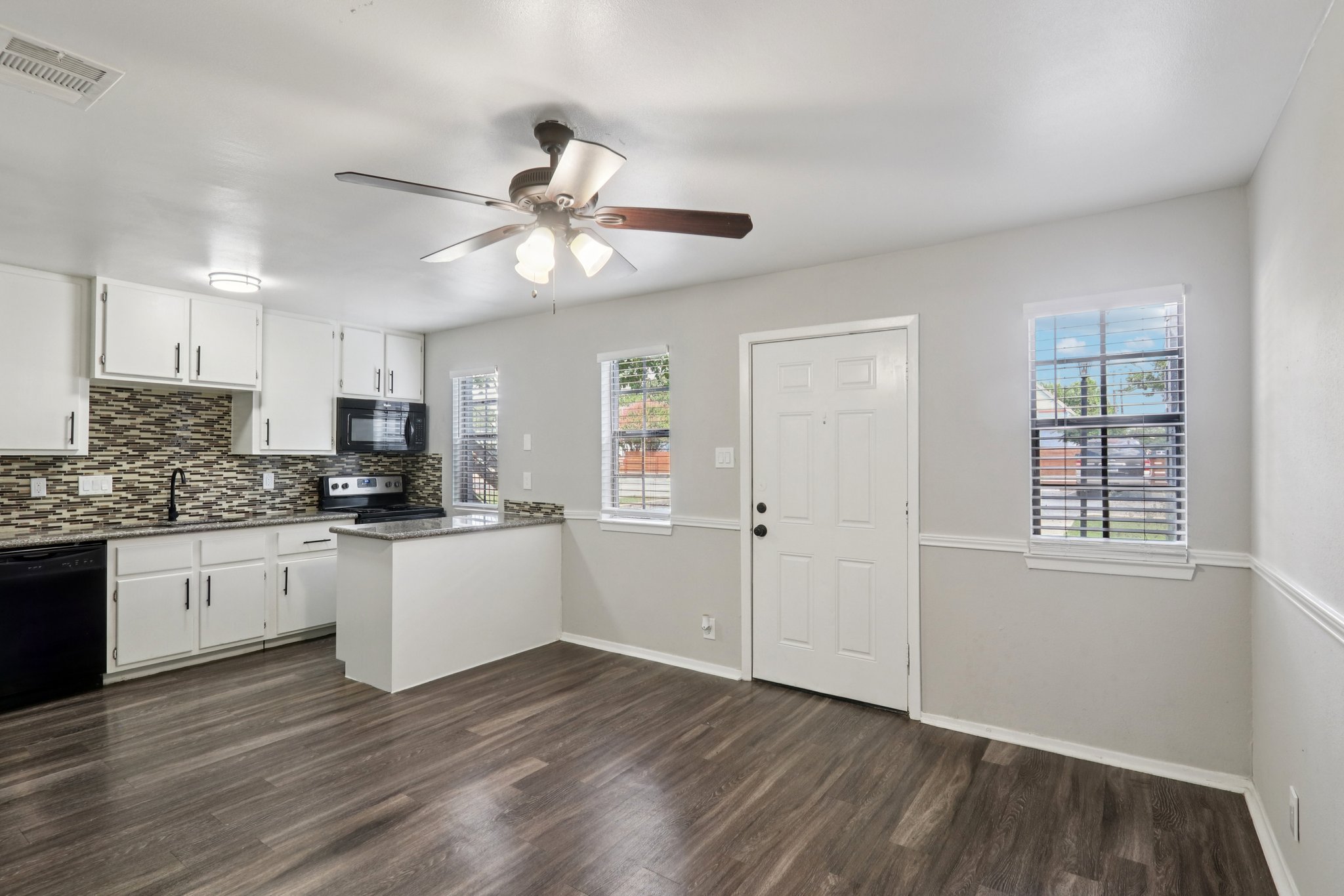 A bright and modern kitchen and living area featuring white cabinets, a black stove, and a ceiling fan. The kitchen has a mosaic tile backsplash and an island with seating. Large windows allow natural light, and there is a door leading outside. The space has wood-like laminate flooring for a cozy feel.