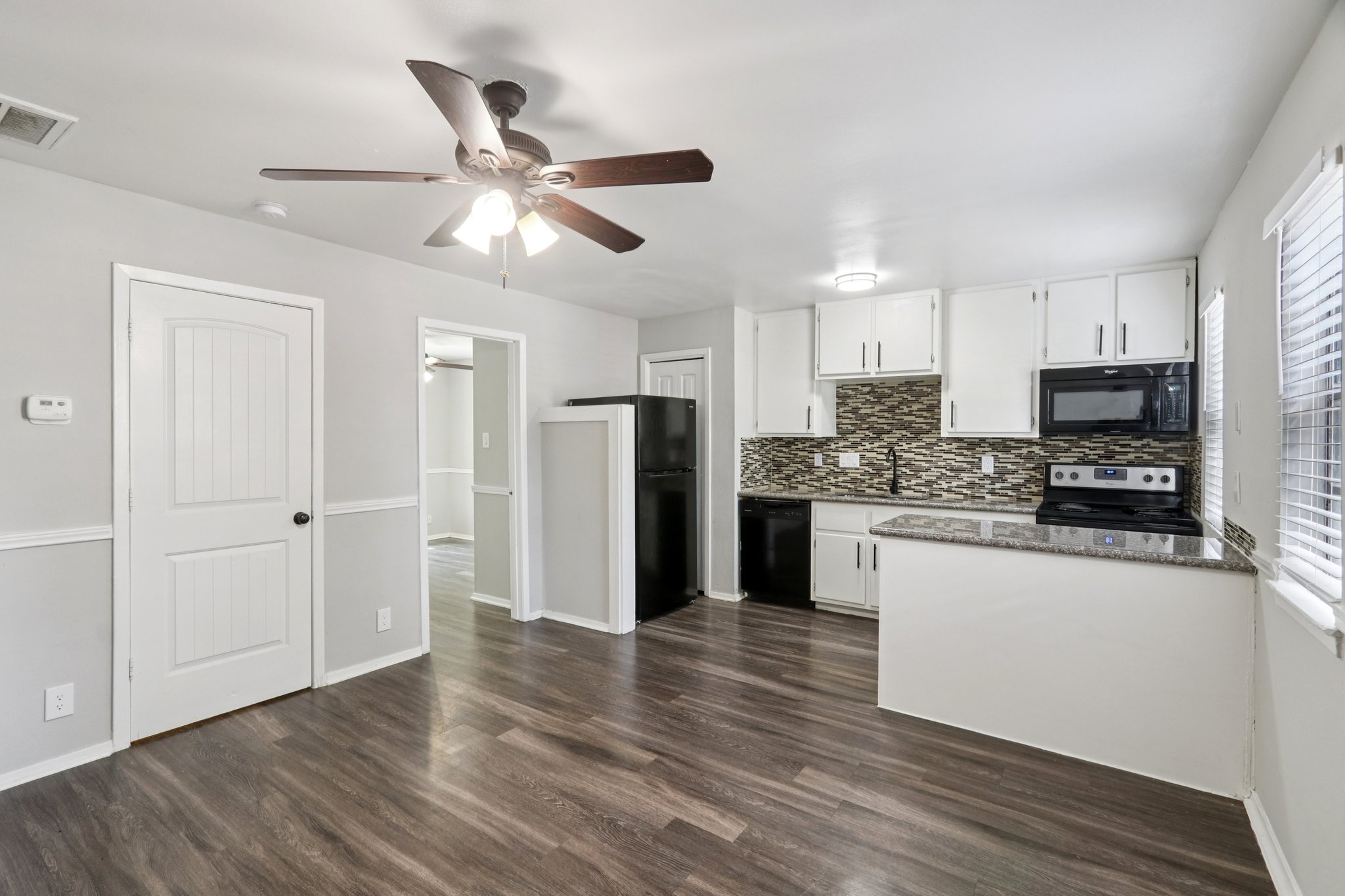 Interior view of a modern kitchen featuring light gray walls, dark wood flooring, black appliances, and a decorative backsplash. The space includes a ceiling fan and white cabinets, with a doorway leading to another room visible in the background. Natural light enters through a window.