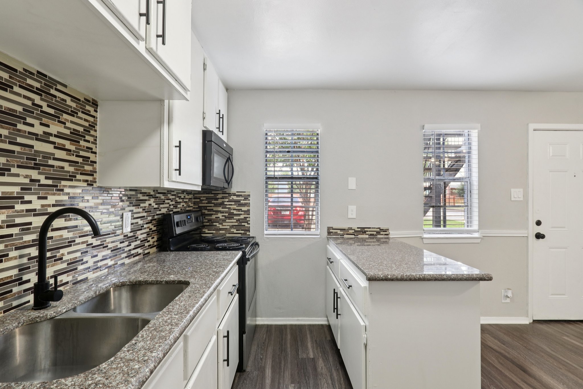 Modern kitchen featuring white cabinetry, a stainless steel sink, and a mosaic tile backsplash. There's a black stove and microwave, with a large countertop and space for breakfast seating. Natural light filters through multiple windows, and a door to the exterior is visible on the right.