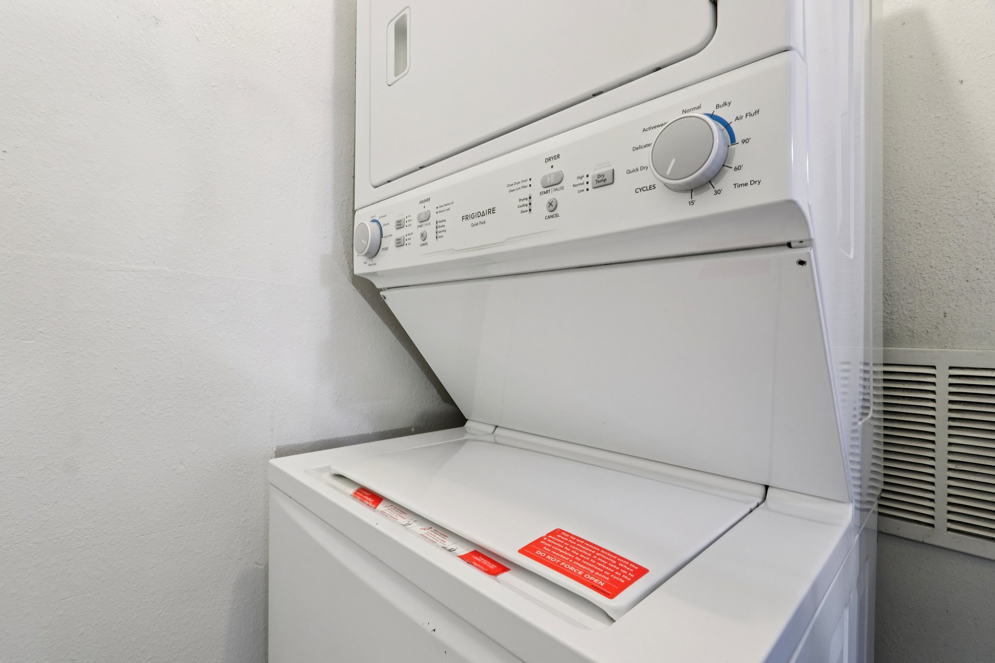 A white stacked washer and dryer unit, featuring control dials and buttons for operation. The dryer is on top, while the washing machine is below. A red warning label is visible on the washing machine's lid. The background is a simple, light-colored wall.