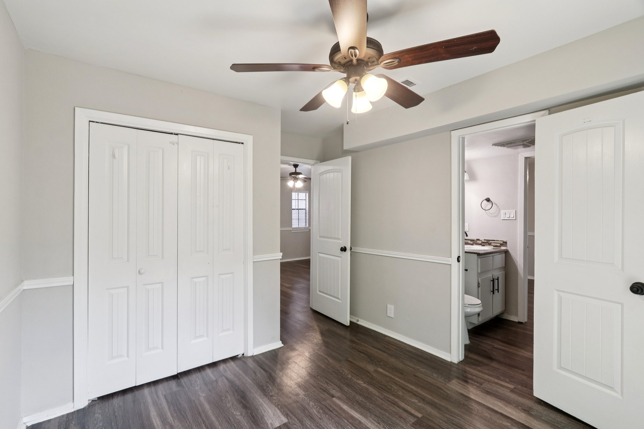 A well-lit room featuring a ceiling fan with wooden blades, light gray walls, and dark laminate flooring. Two white double closet doors are visible on the left, and a doorway on the right leads to a bathroom. Natural light enters from a window, enhancing the spacious feel of the area.