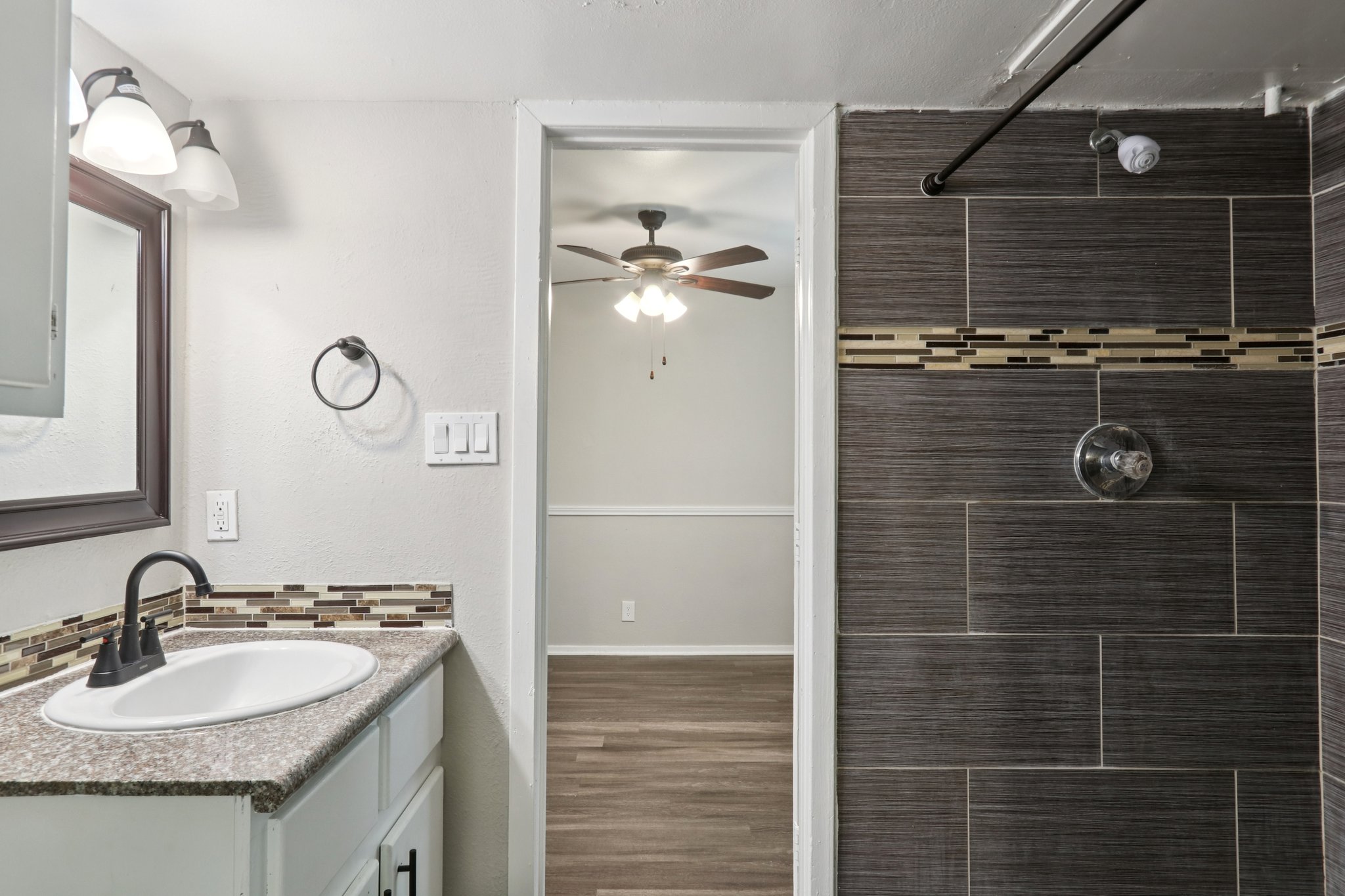 A modern bathroom featuring a sink with a granite countertop, a mirror, and a shower area with dark tiled walls. A door leads to a light-colored room with a ceiling fan and wood-like flooring. The walls are painted in neutral tones, giving a clean and contemporary look.