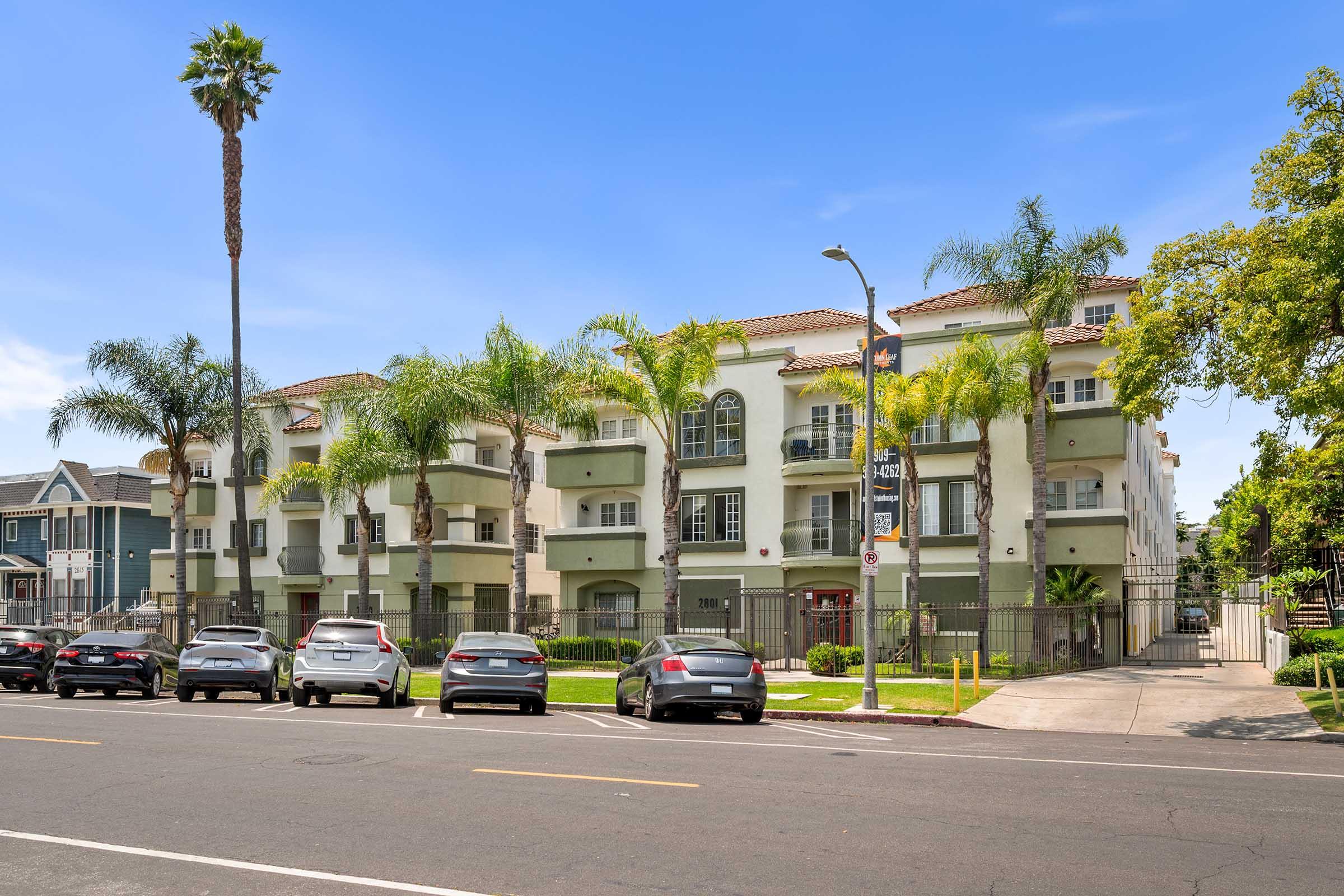 A modern apartment building with a light-colored facade and palm trees in front. Several cars are parked along the street in front of the building, and there is a clear blue sky overhead. The entrance is gated, providing additional security for the residents.
