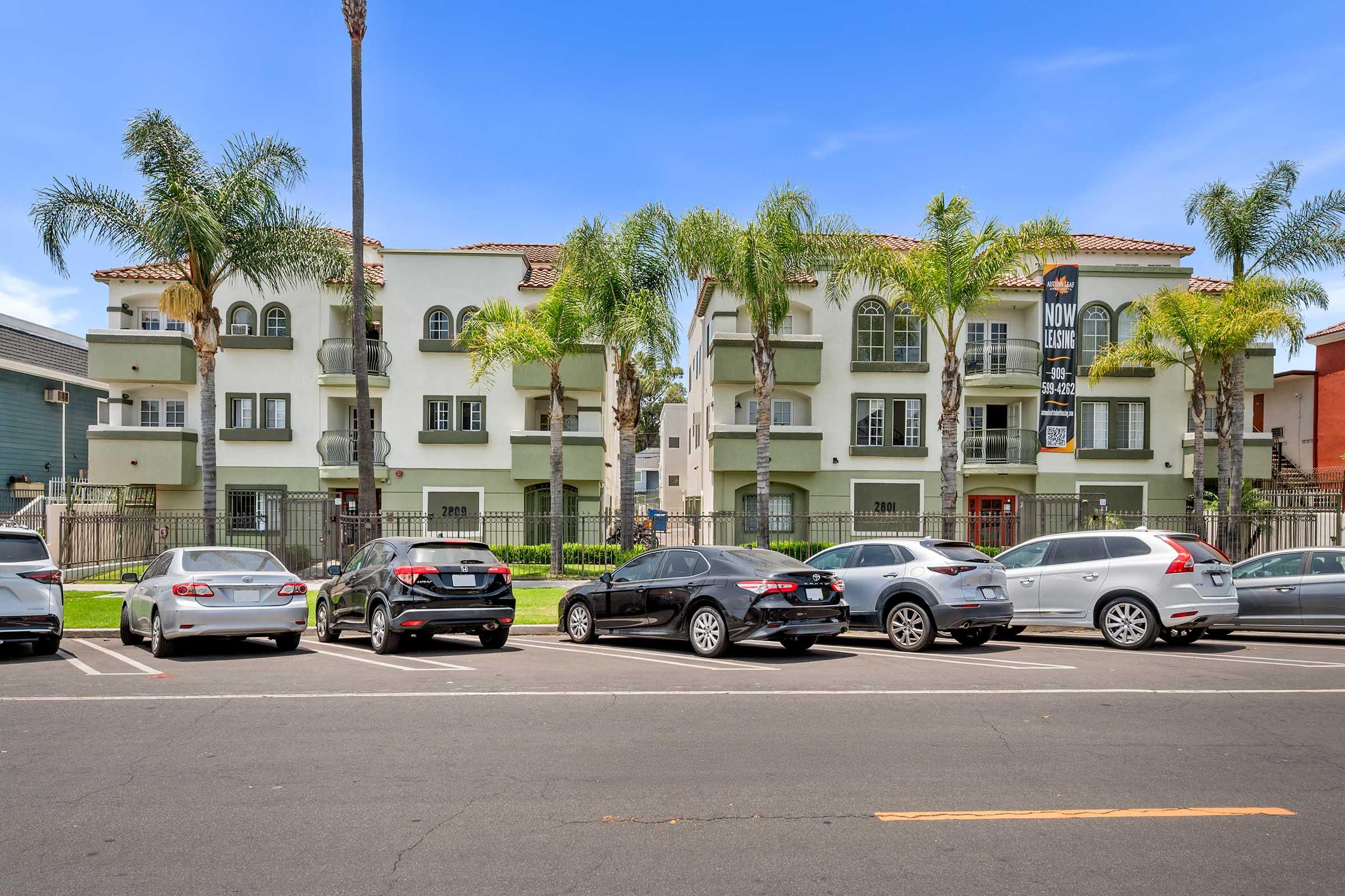A residential apartment building with three stories, featuring palm trees in the front and a well-maintained lawn. Several cars are parked along the street in front of the building, which has a sign advertising availability. The sky is clear and blue, creating a sunny atmosphere.