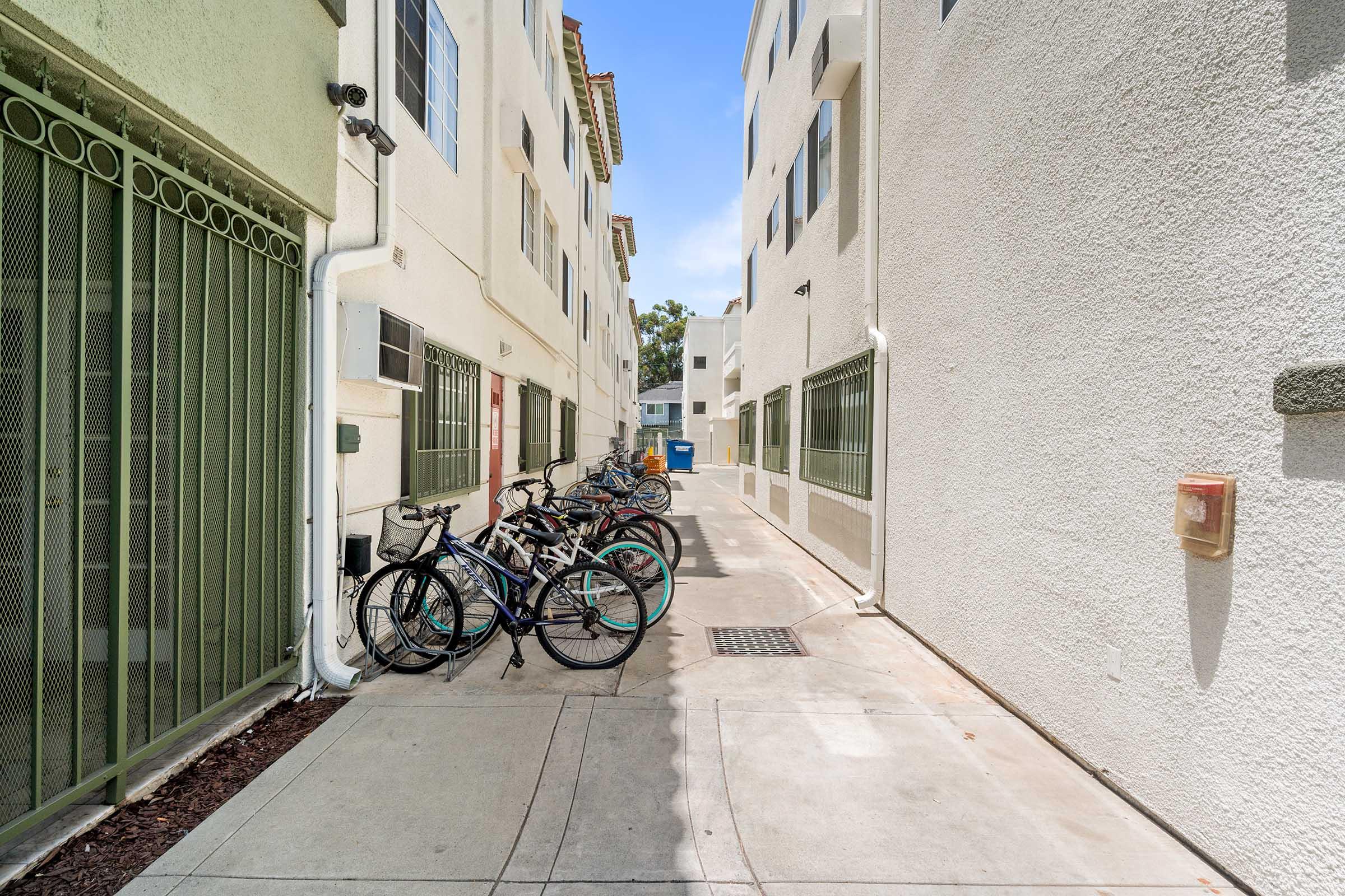 Narrow alleyway between residential buildings with several bicycles parked along one side. The walls are painted in light colors, and there are windows on both sides. Bright blue sky is visible at the end of the alley.