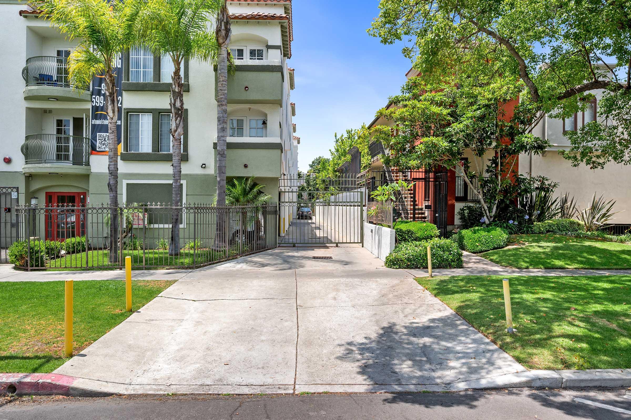 A view of a residential street featuring two apartment buildings flanking a gated entrance. The left building is green with balconies, while the right is orange with lush landscaping. Palm trees and green shrubs add to the scene, with a clear blue sky overhead. A paved driveway leads to the gate.