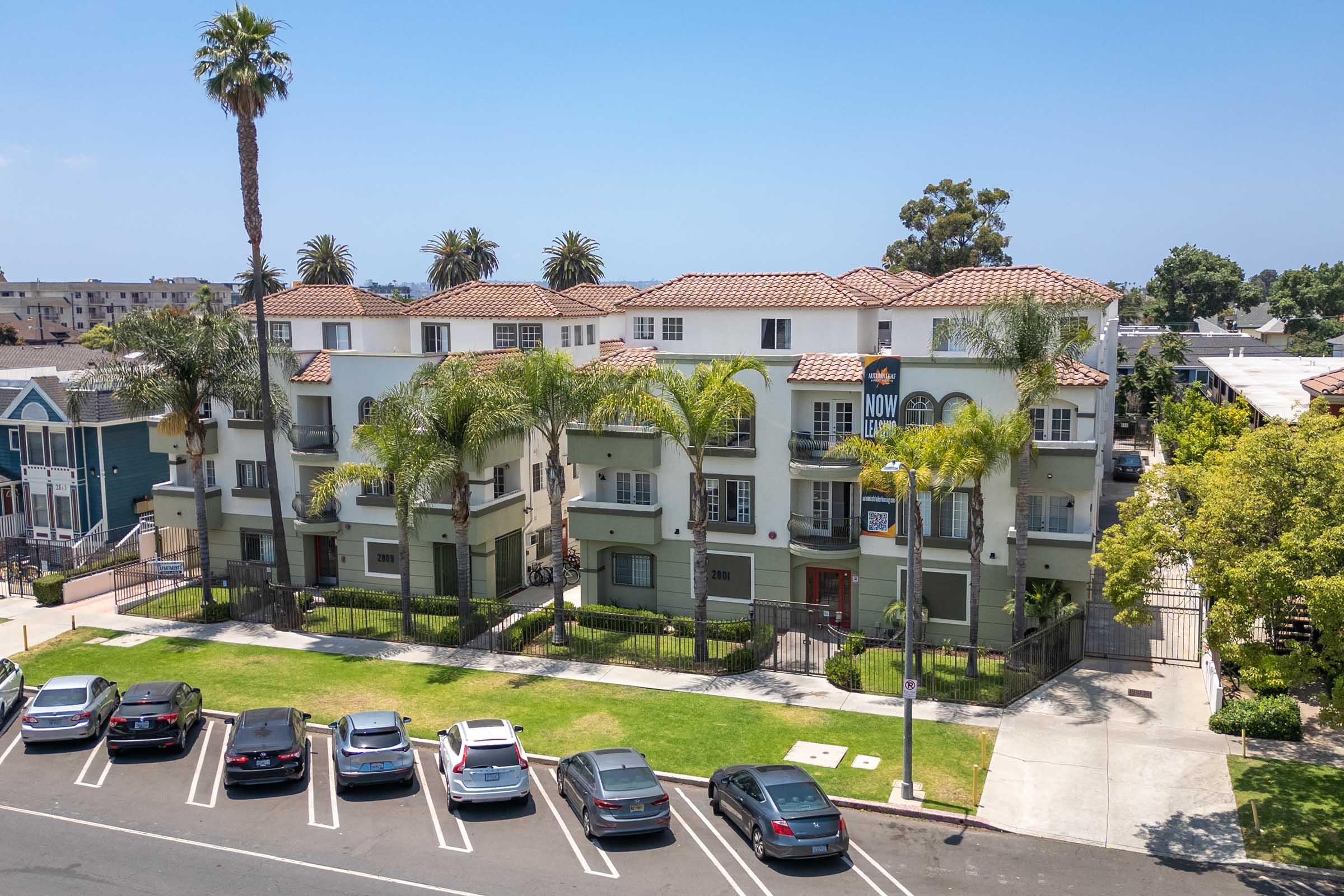 Aerial view of a multi-story apartment building surrounded by palm trees and greenery. The building features a light green exterior with a tile roof and is enclosed by a fence. Several parked cars are visible in front, along with a lawn area. Blue sky in the background.