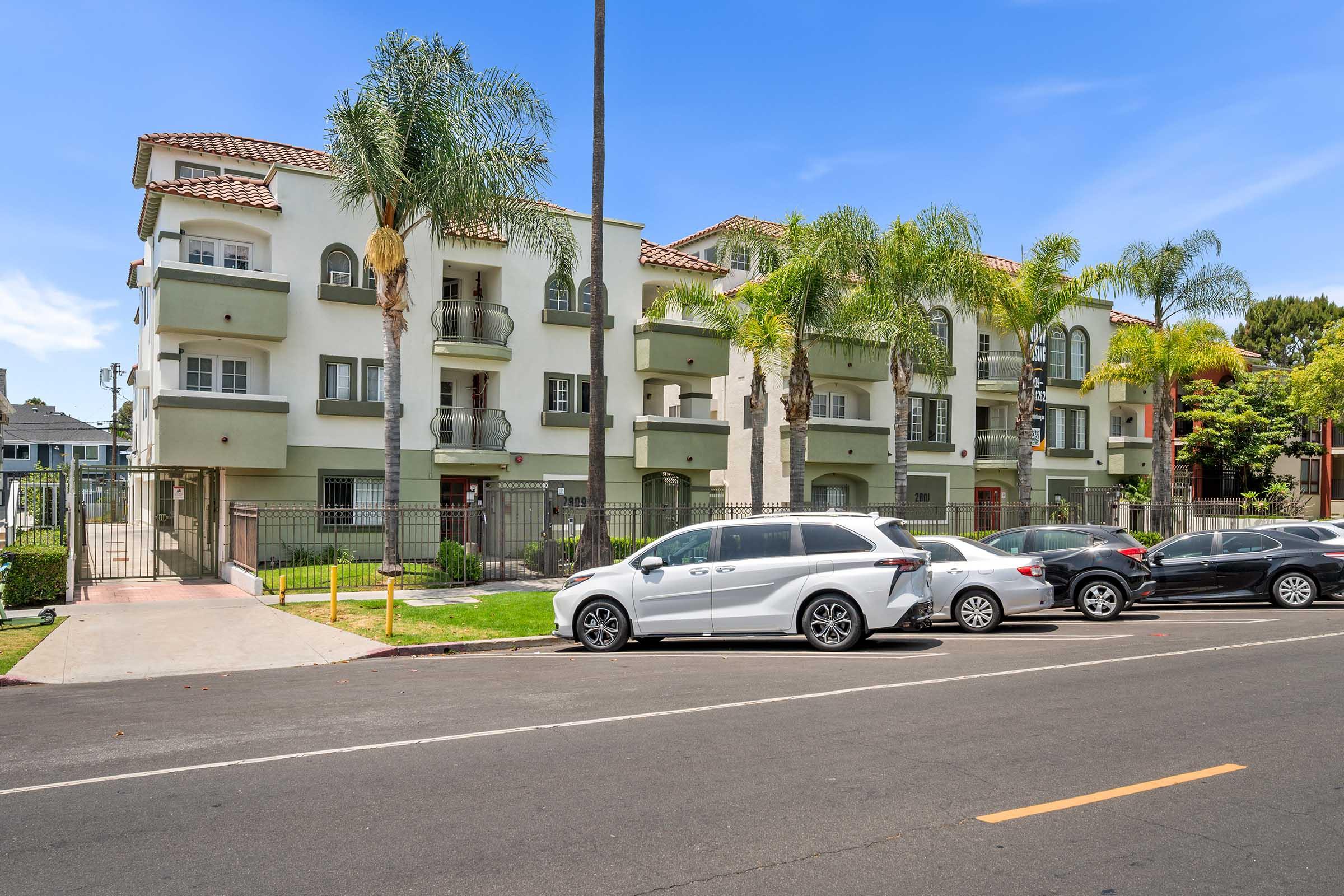 A modern apartment building with a light-colored facade, featuring balconies and palm trees in the foreground. A paved road runs alongside, with several parked cars. The sky is clear and blue, adding to the sunny atmosphere of the scene.