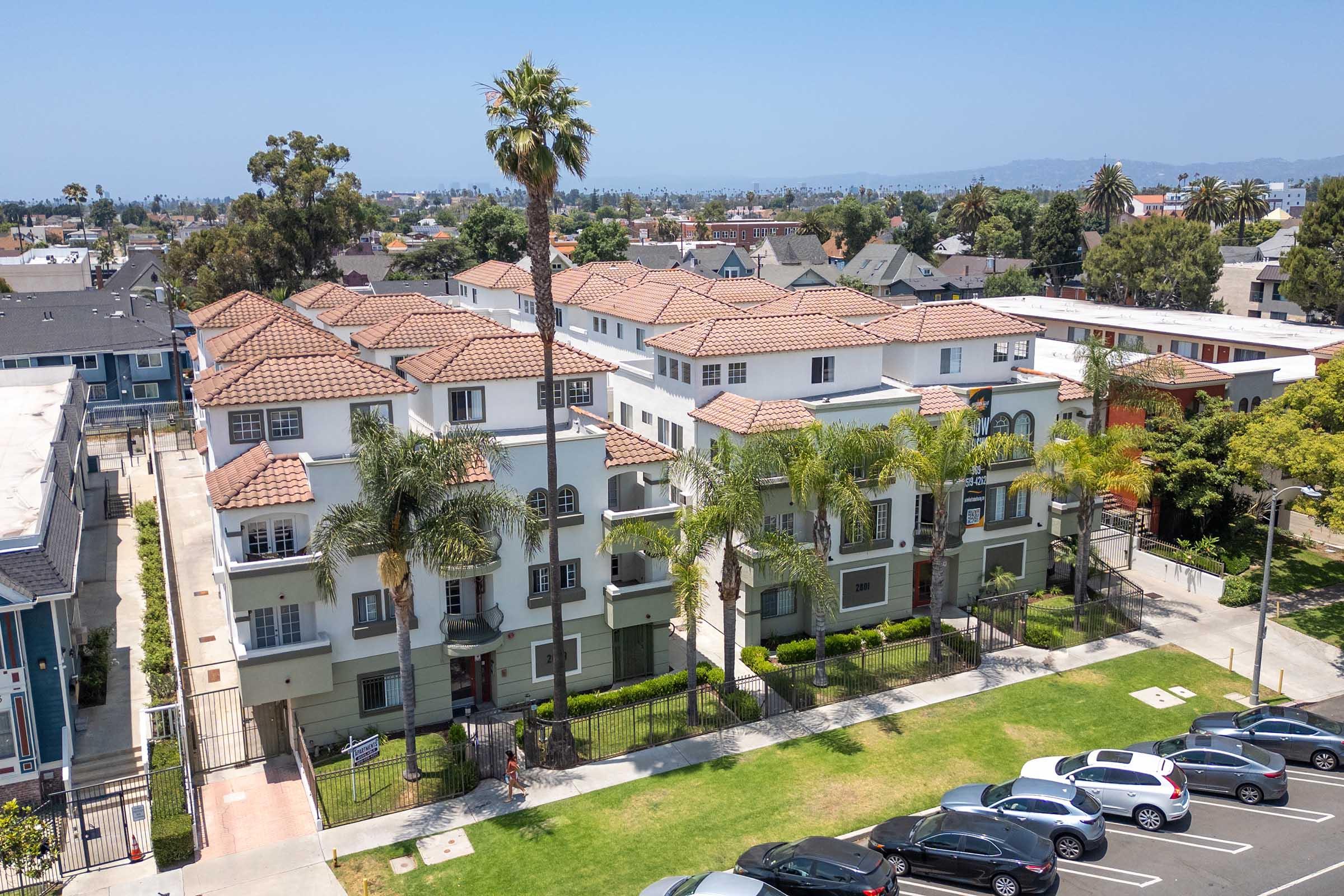 Aerial view of a residential complex with multiple two-story buildings featuring tile roofs, surrounded by palm trees and green lawns. A parking area with several cars is visible in the foreground, and the distant cityscape can be seen in the background under a clear blue sky.