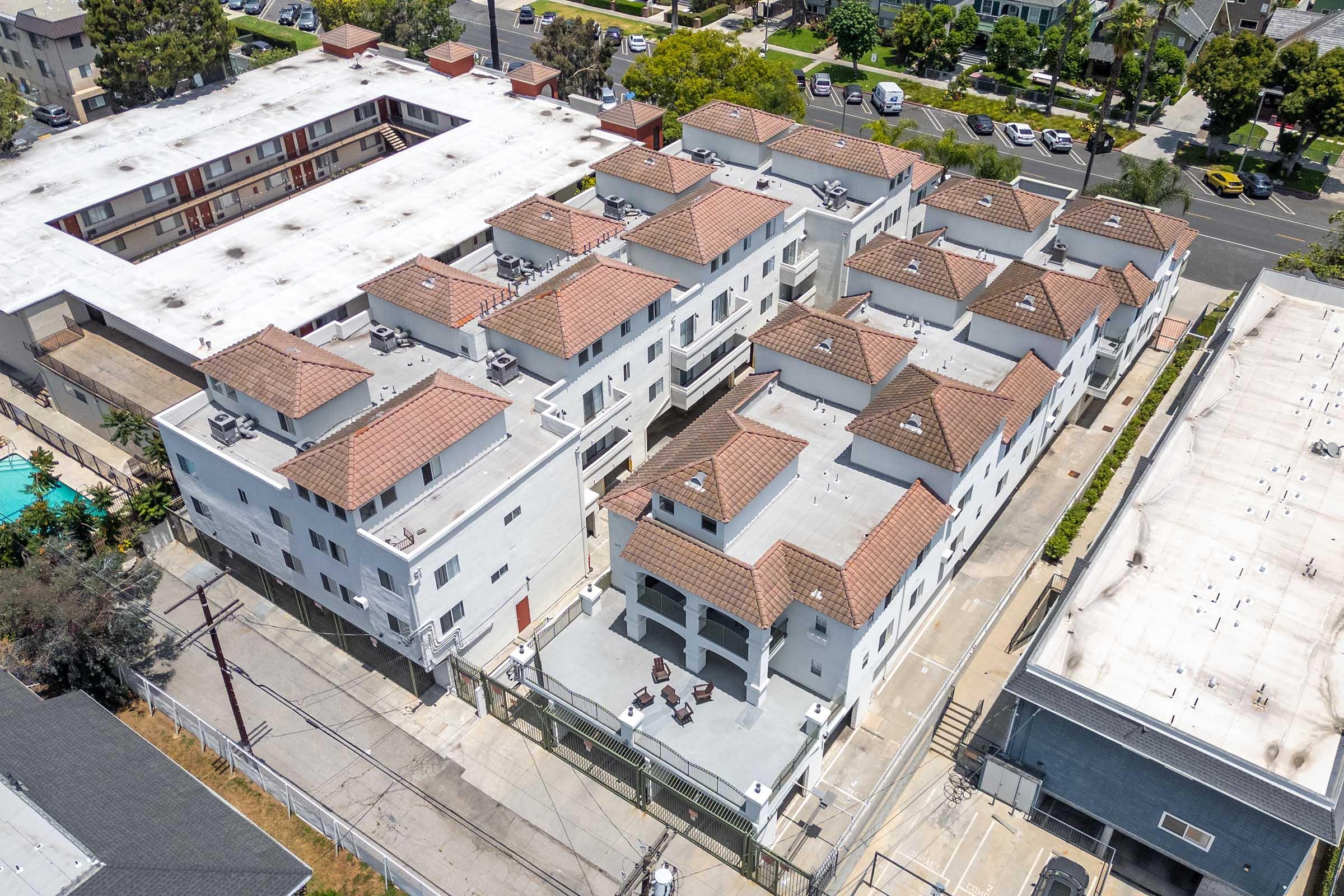 Aerial view of a residential complex featuring white buildings with red-tiled roofs, surrounded by green trees and parked cars. The layout includes multiple units, some with outdoor seating, as well as a larger building in the background. The scene captures a mix of urban and residential environments.