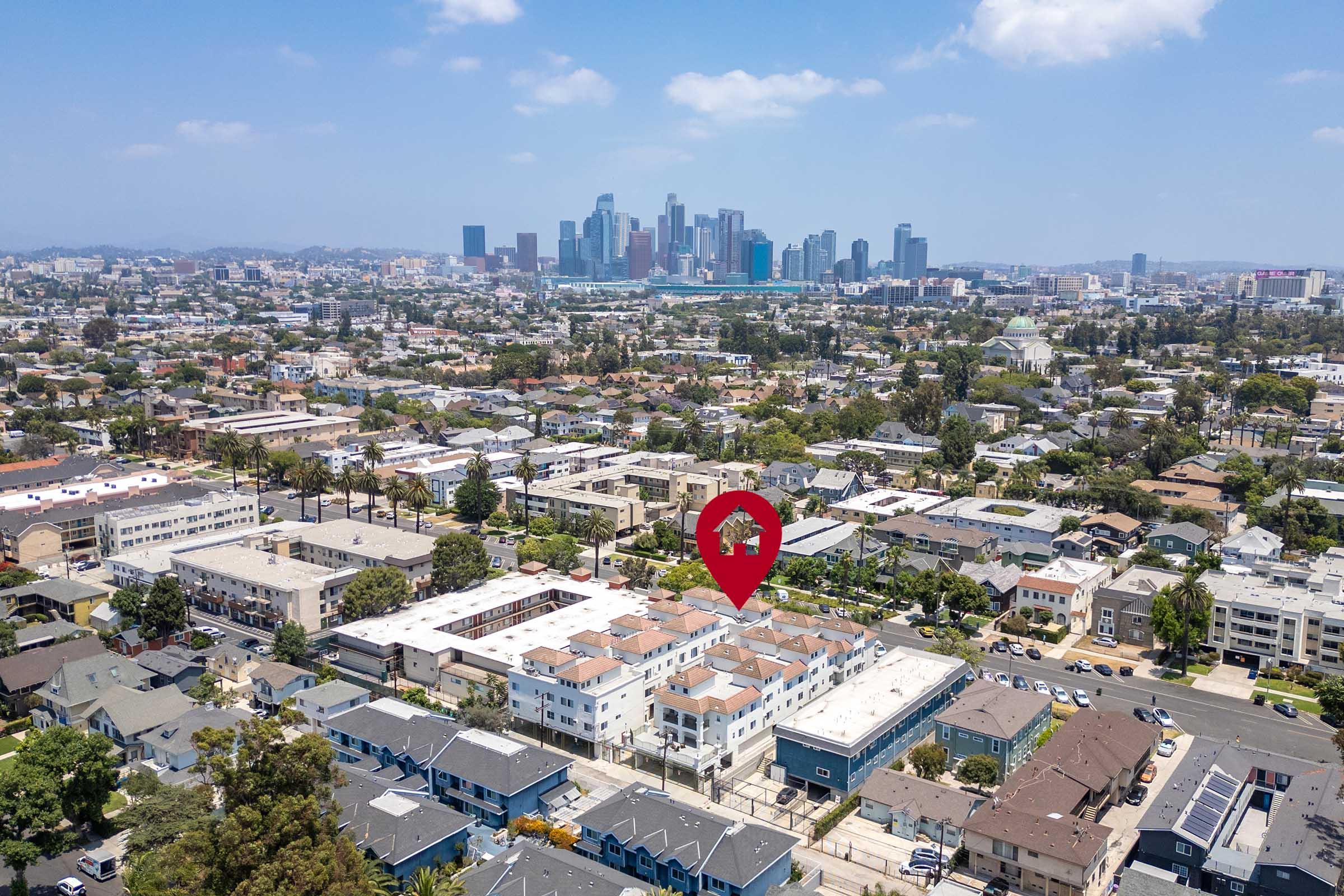 Aerial view of a cityscape featuring a cluster of residential buildings in the foreground with a red pin marking a specific location. In the background, the skyline of a bustling downtown area with skyscrapers under a partly cloudy sky.