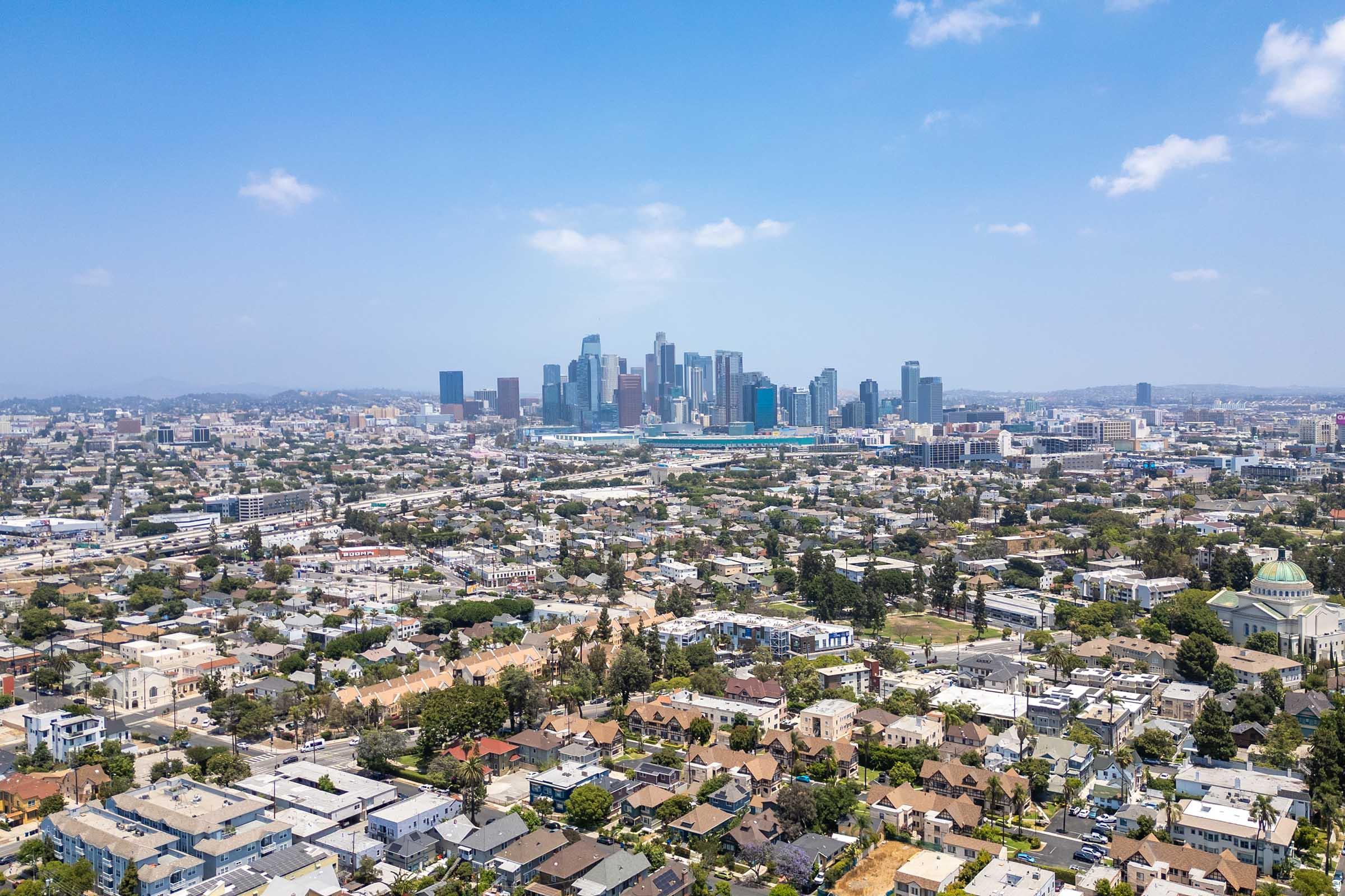 Aerial view of a cityscape featuring downtown Los Angeles with tall skyscrapers in the background. In the foreground, a mix of residential neighborhoods and green spaces is visible. The sky is clear with few clouds, showcasing a vibrant urban setting.