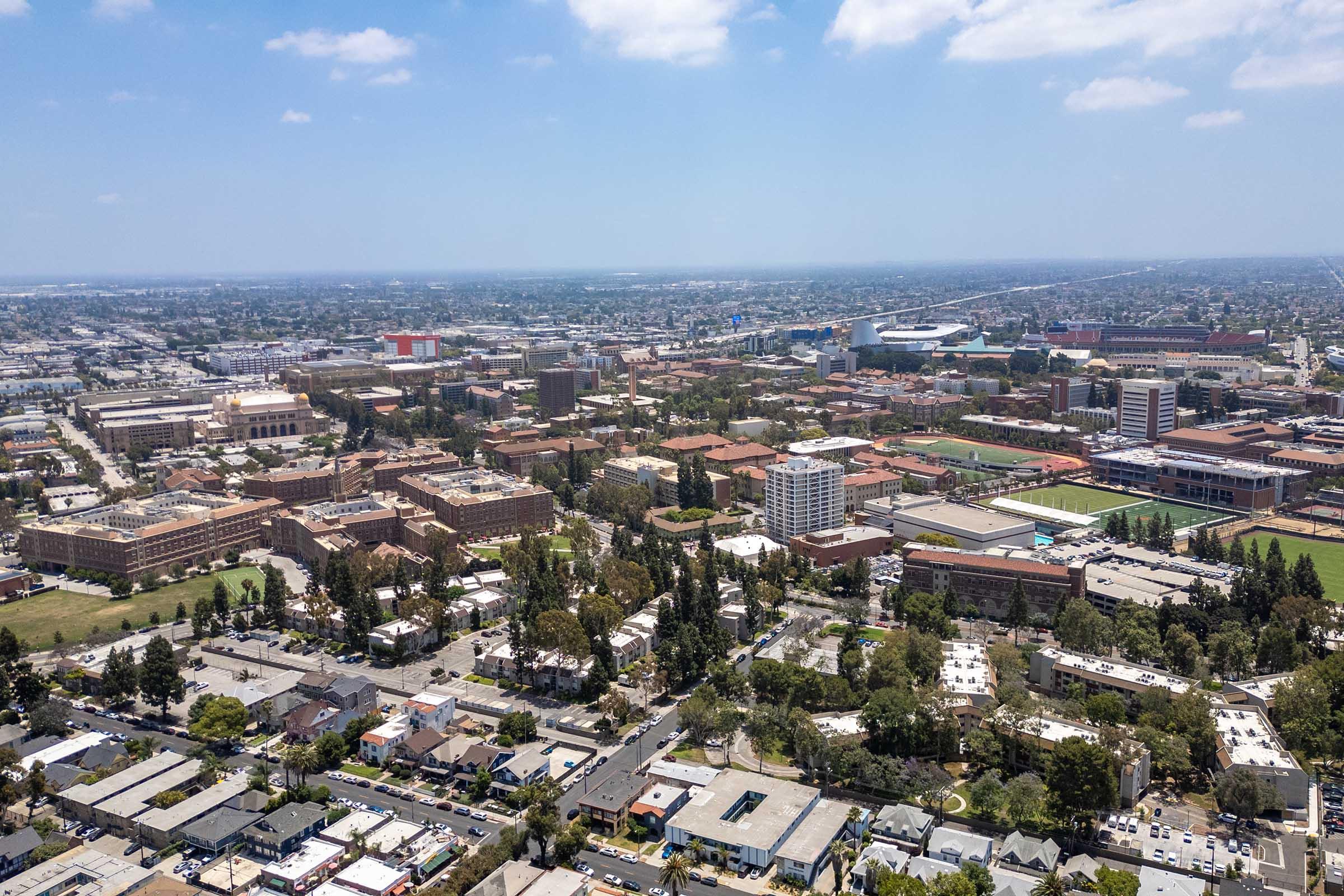 Aerial view of a university campus showing a mix of buildings, green spaces, and sports facilities. The surrounding area includes residential neighborhoods and roads, with a clear sky above. The layout showcases the scale and architectural diversity of the campus environment.