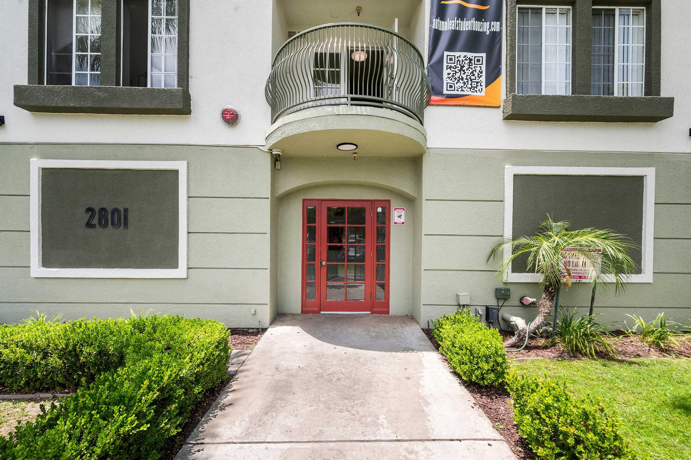 Entrance of a building featuring a set of double doors in red, surrounded by light green walls. The address "2801" is displayed on the wall. There are neatly trimmed bushes on both sides of the walkway leading up to the doors, and a small palm tree is visible to the right. A banner hangs above with a QR code.