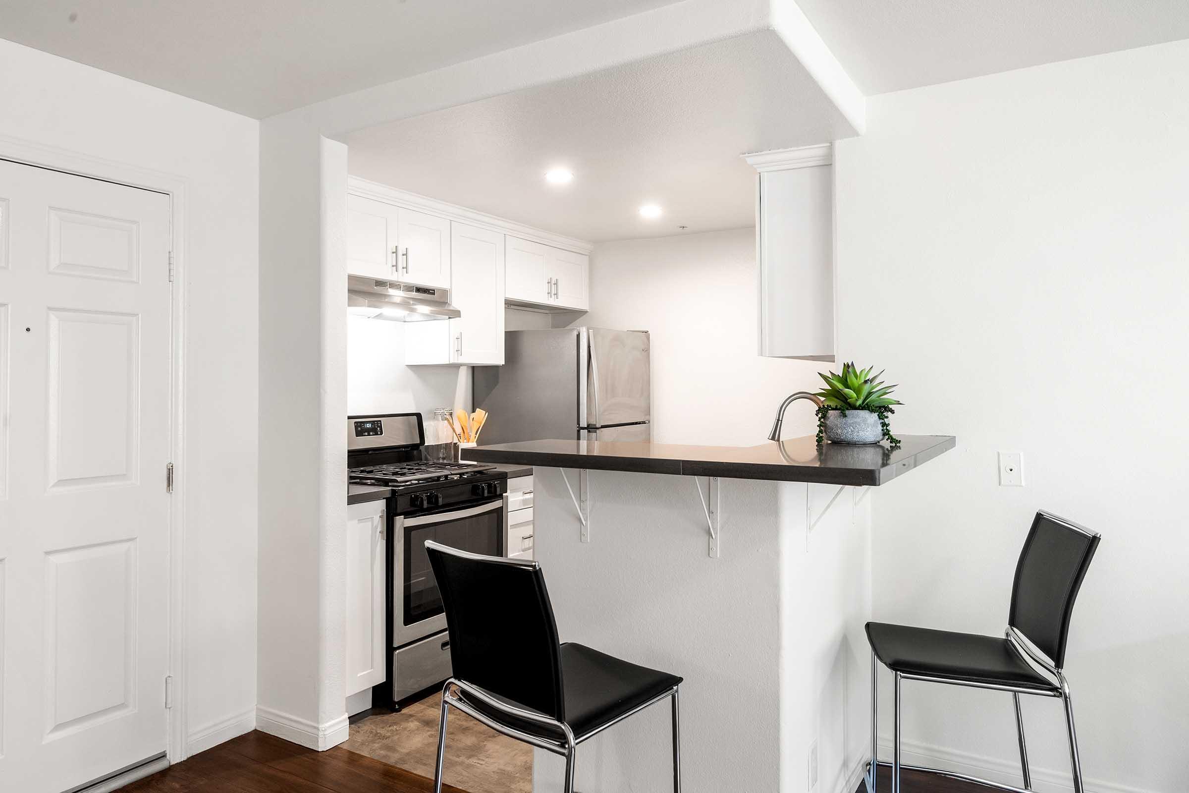 A modern kitchen area featuring a black countertop with a small overhang, stainless steel appliances, and white cabinetry. Two black chairs are situated at the countertop. The room has a light-colored wall and hardwood flooring, creating a bright, inviting atmosphere. A door is visible on the left side.