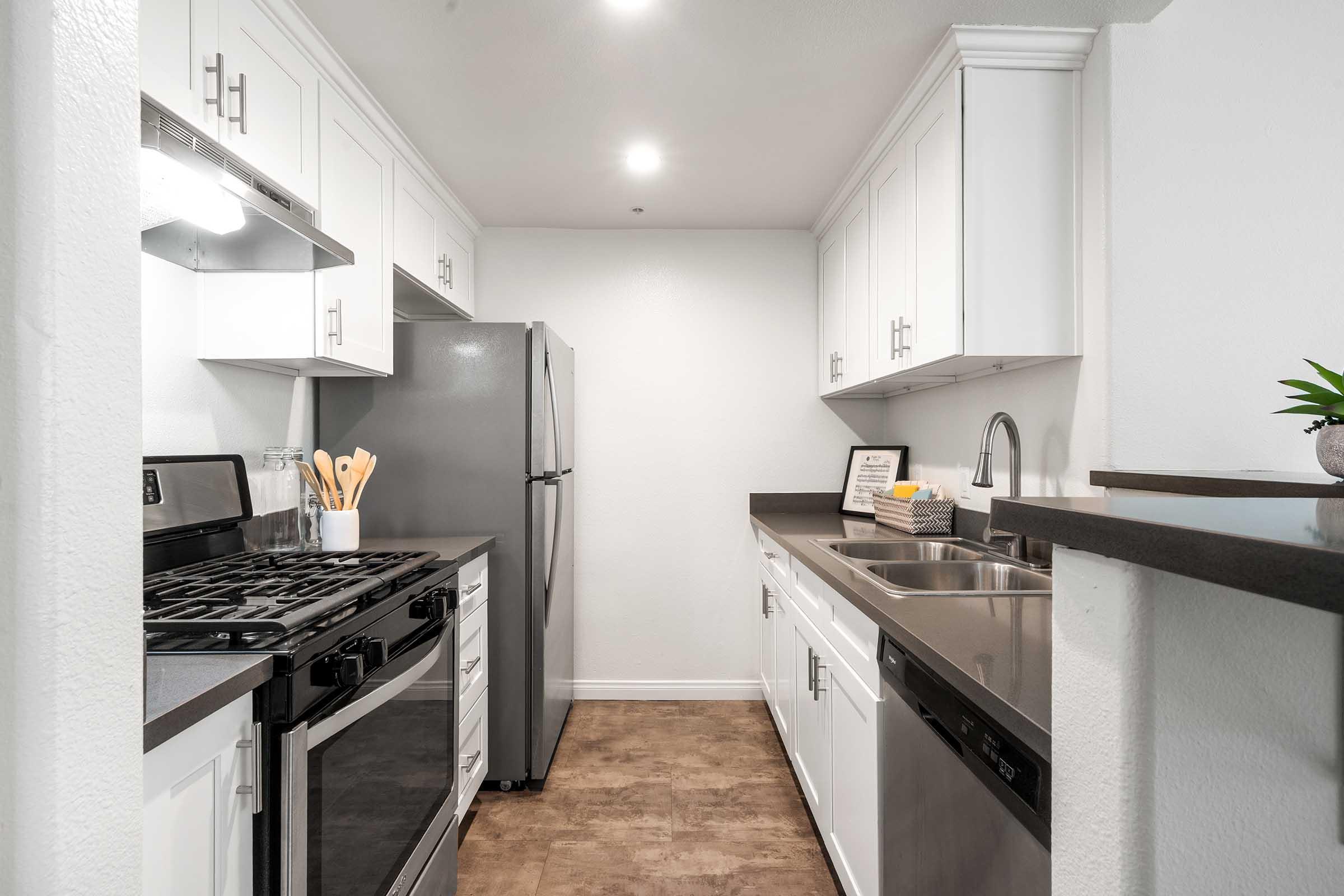 Modern kitchen featuring white cabinets, a stainless steel refrigerator, a gas stove, and a double sink. The countertop is dark, and there are kitchen utensils on display. Bright overhead lighting illuminates the space, creating a clean and inviting atmosphere.