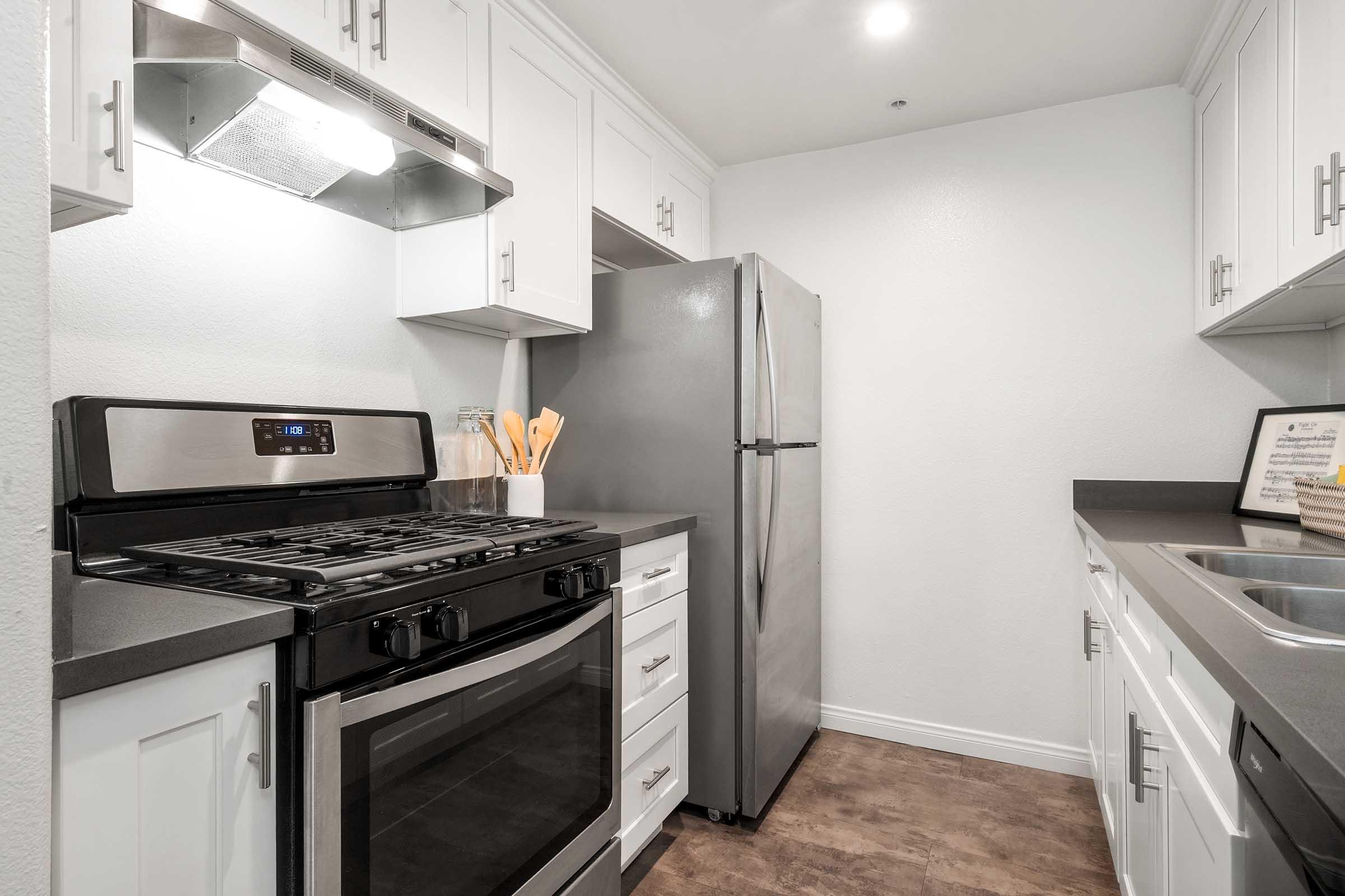A modern kitchen featuring white cabinets, a stainless steel refrigerator, a stove with an oven, and a sink. The countertops are dark gray, and the floor has a wood-like appearance. Cooking utensils are displayed near the stove, with bright overhead lighting illuminating the space.